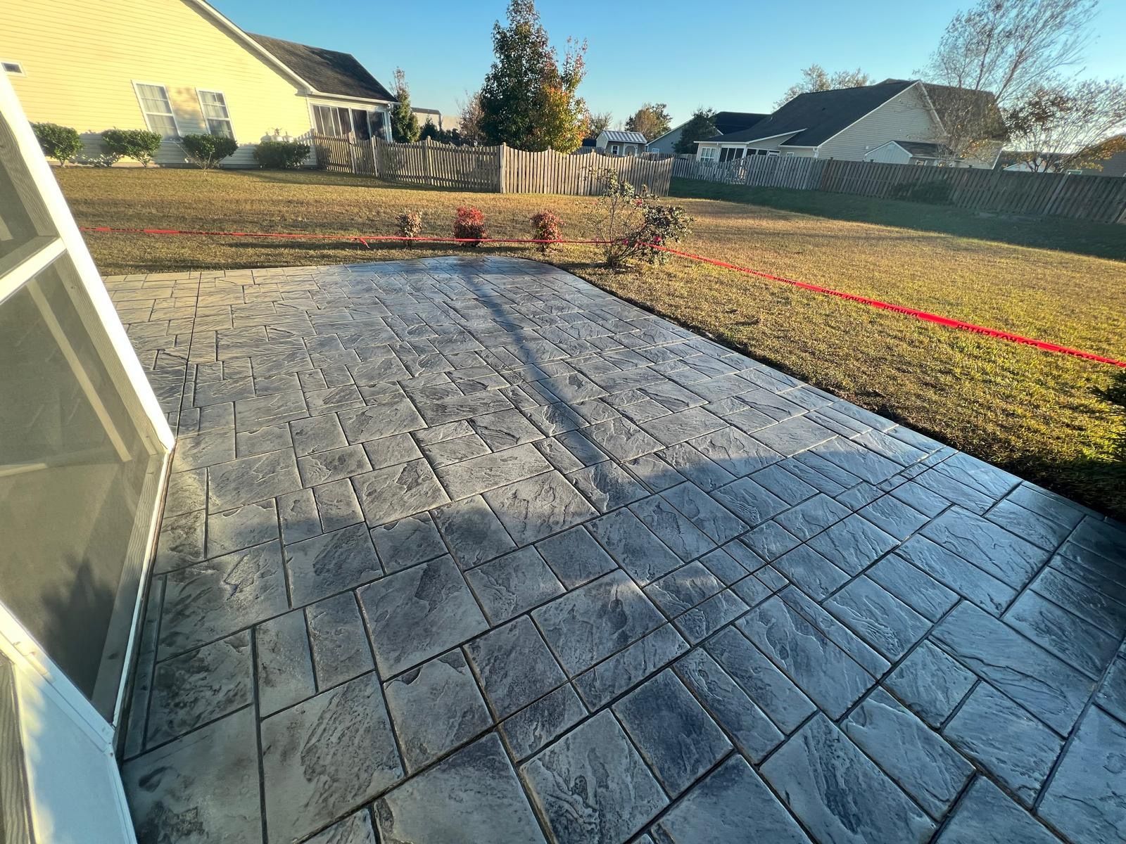 Backyard patio with gray stamped concrete, grassy yard, and houses in the background under a sunny sky