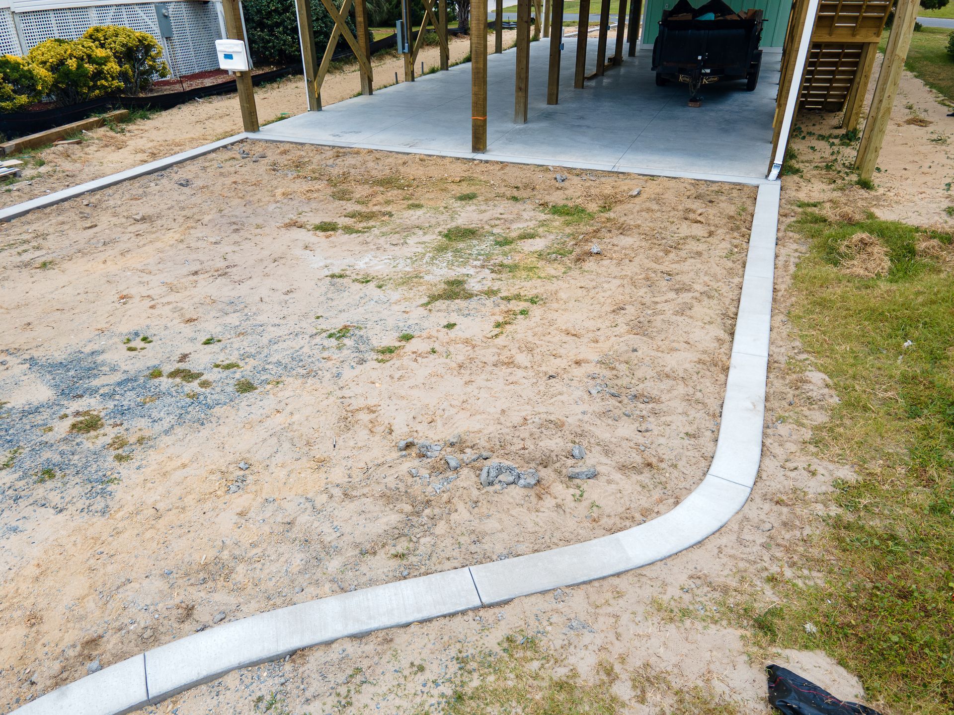 Concrete driveway beside a sandy yard, with a carport and parked vehicle in the background.