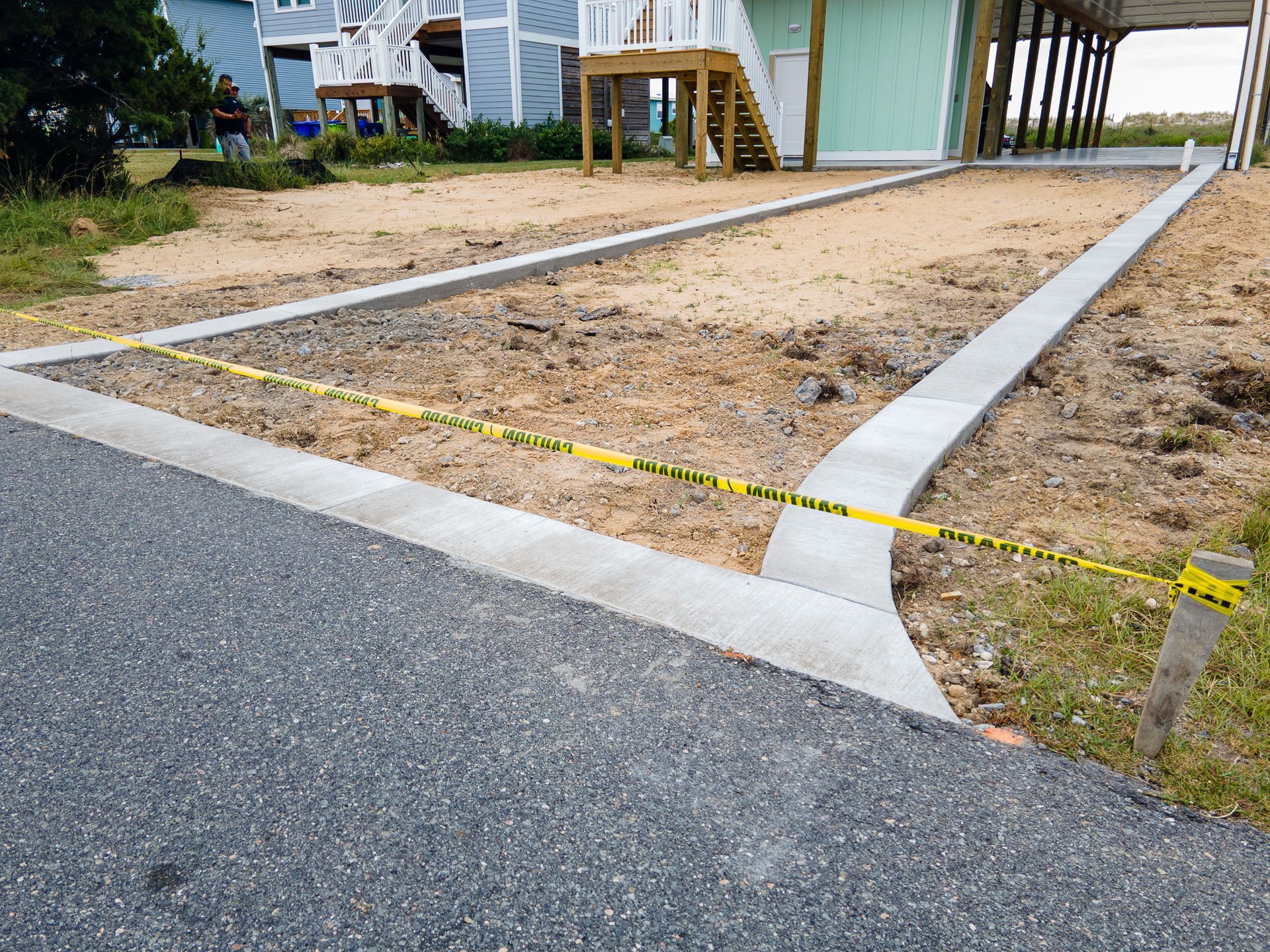 Empty gravel lot beside a paved road, marked with yellow tape and bordered by a house on stilts.