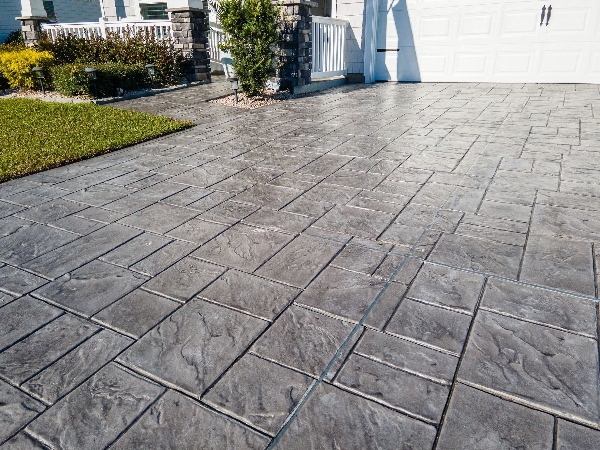 Stamped concrete driveway leading to a white garage beside a green lawn and shrubs