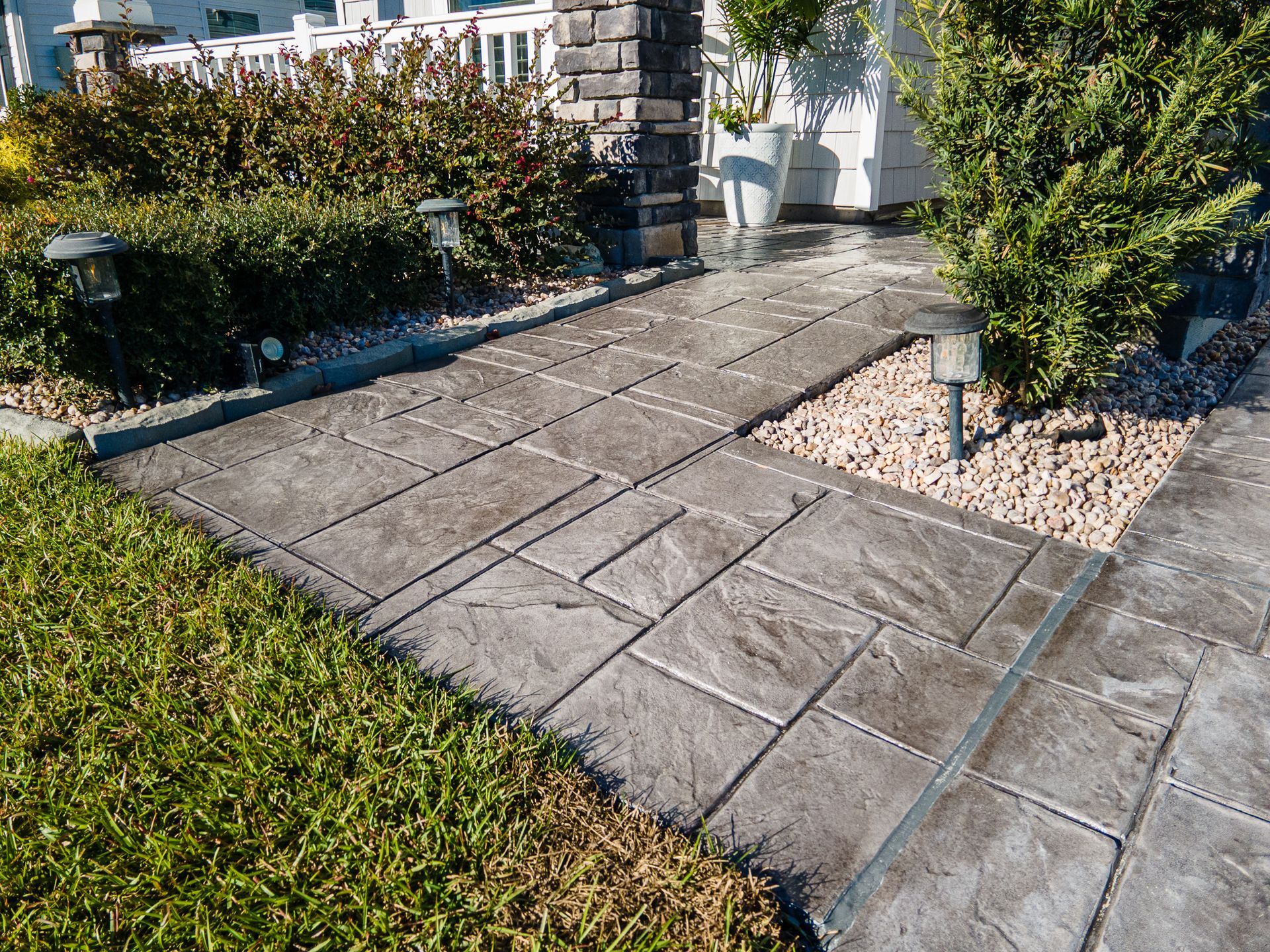 Stone walkway beside trimmed hedges and a porch, with a patch of grass in the foreground