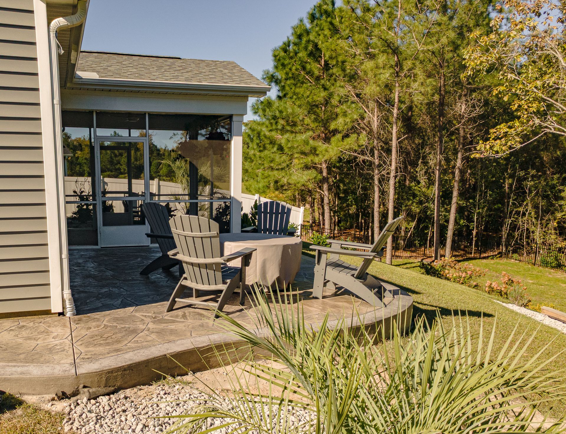 Patio with outdoor chairs and table beside a house, overlooking a sunny backyard with trees and lawn