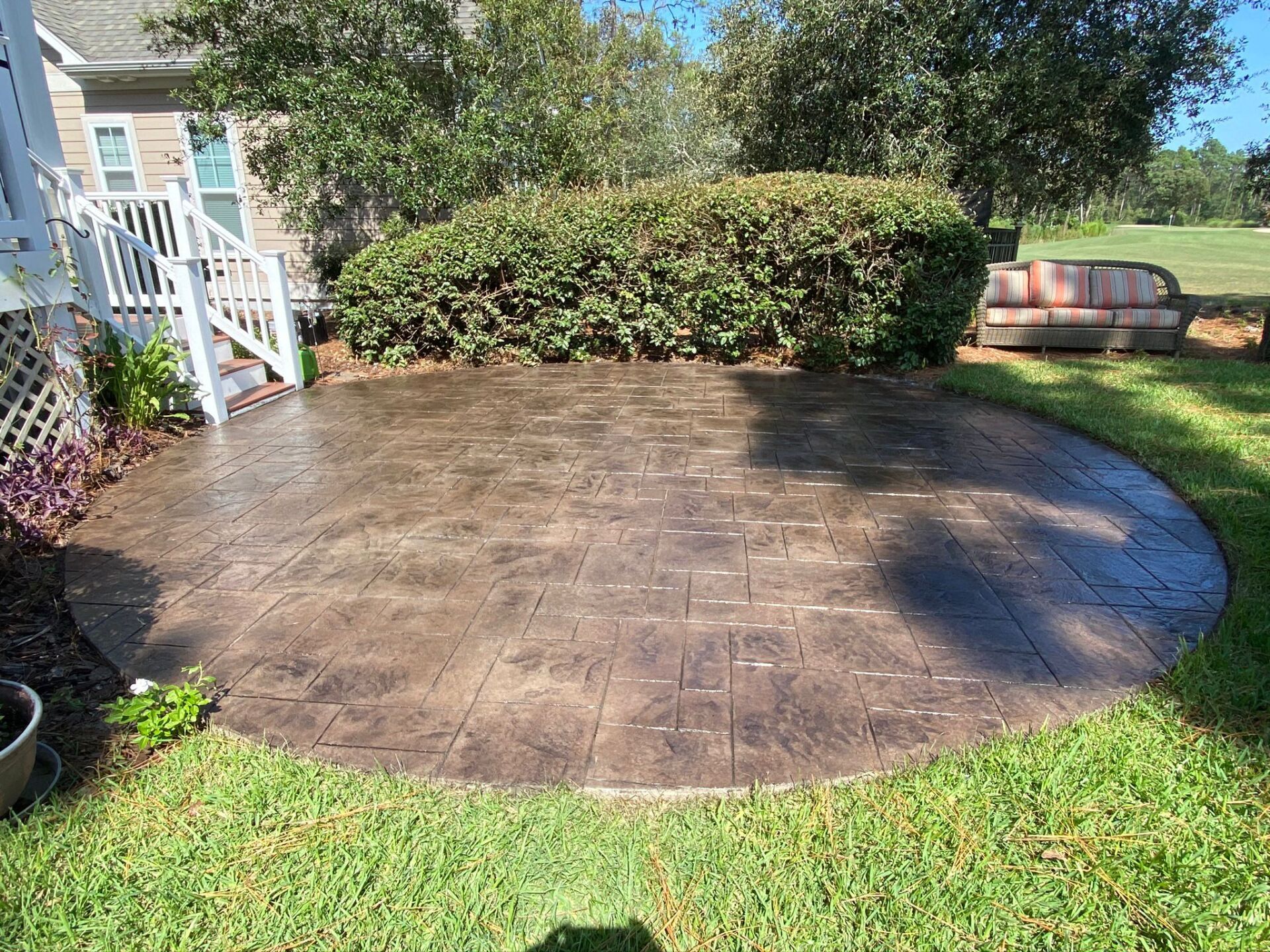 Circular stamped-concrete patio in a backyard with shrubs, lawn, and a house deck nearby
