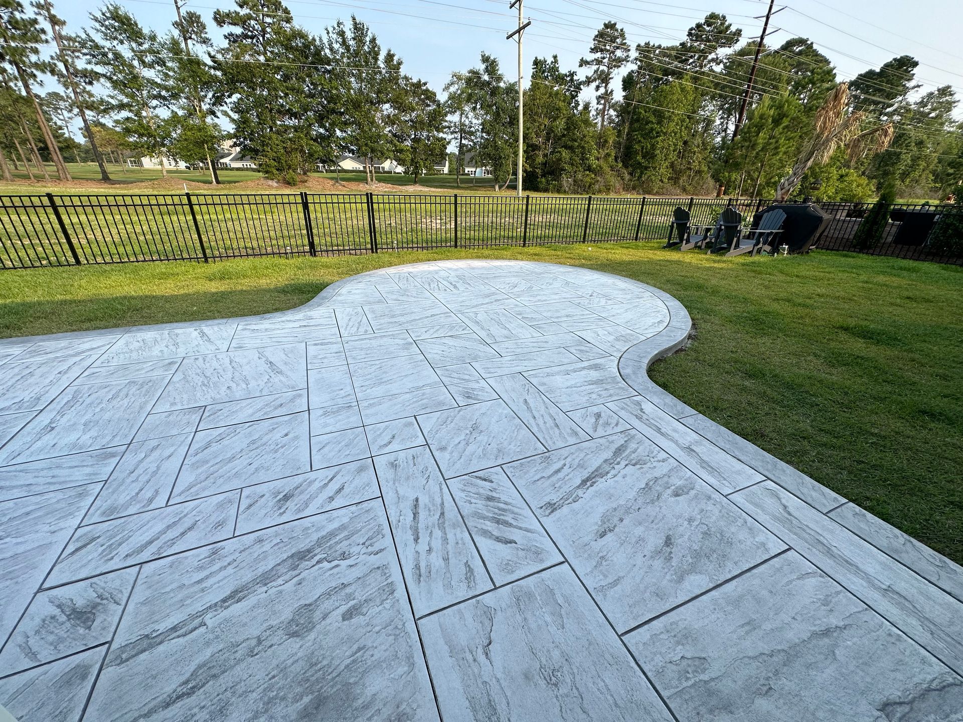 Freshly finished light-gray stamped concrete patio beside a grassy yard and fence under a sunny sky