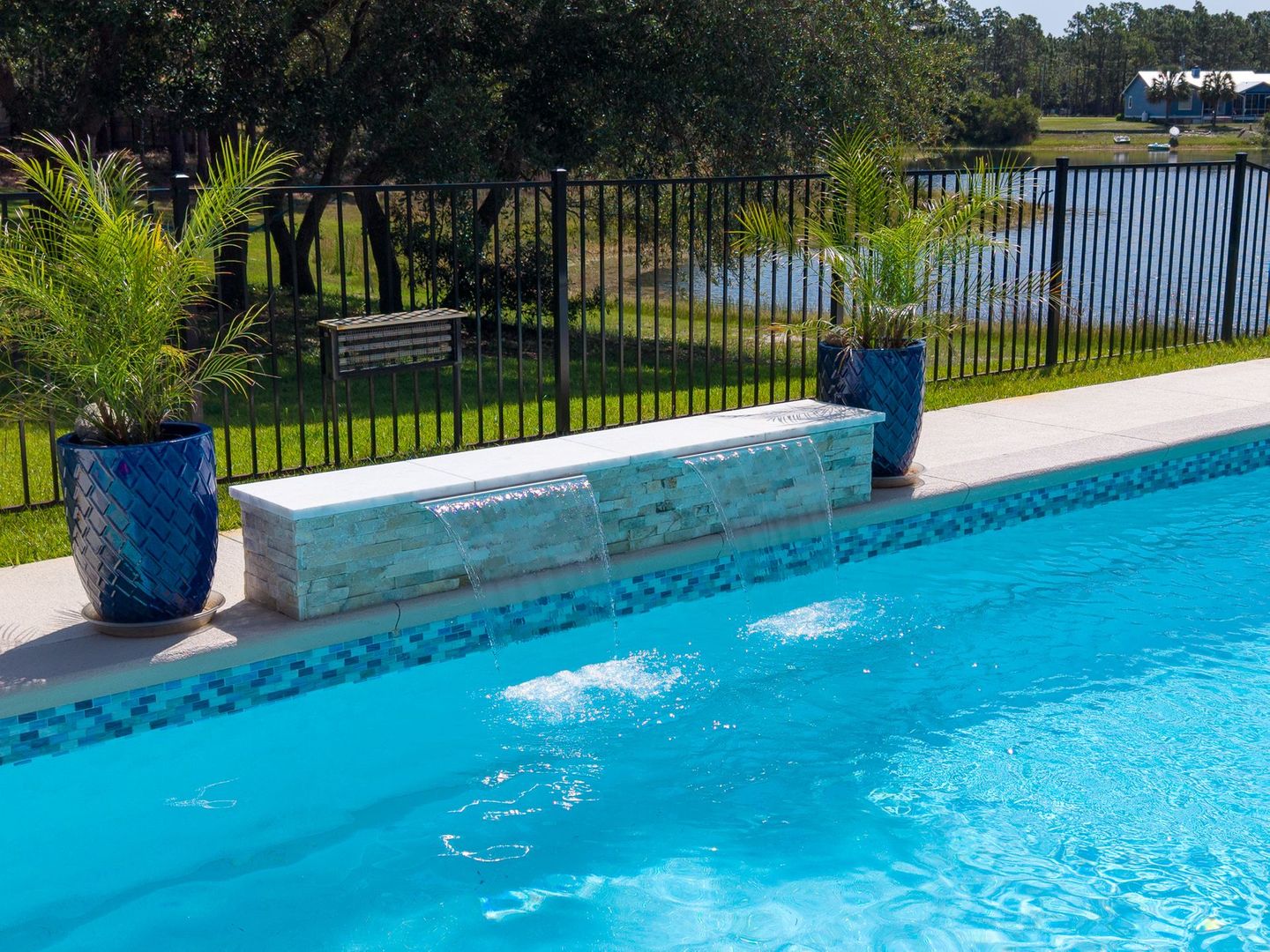 Swimming pool with blue water, stone bench, and potted plants by a black fence outdoors