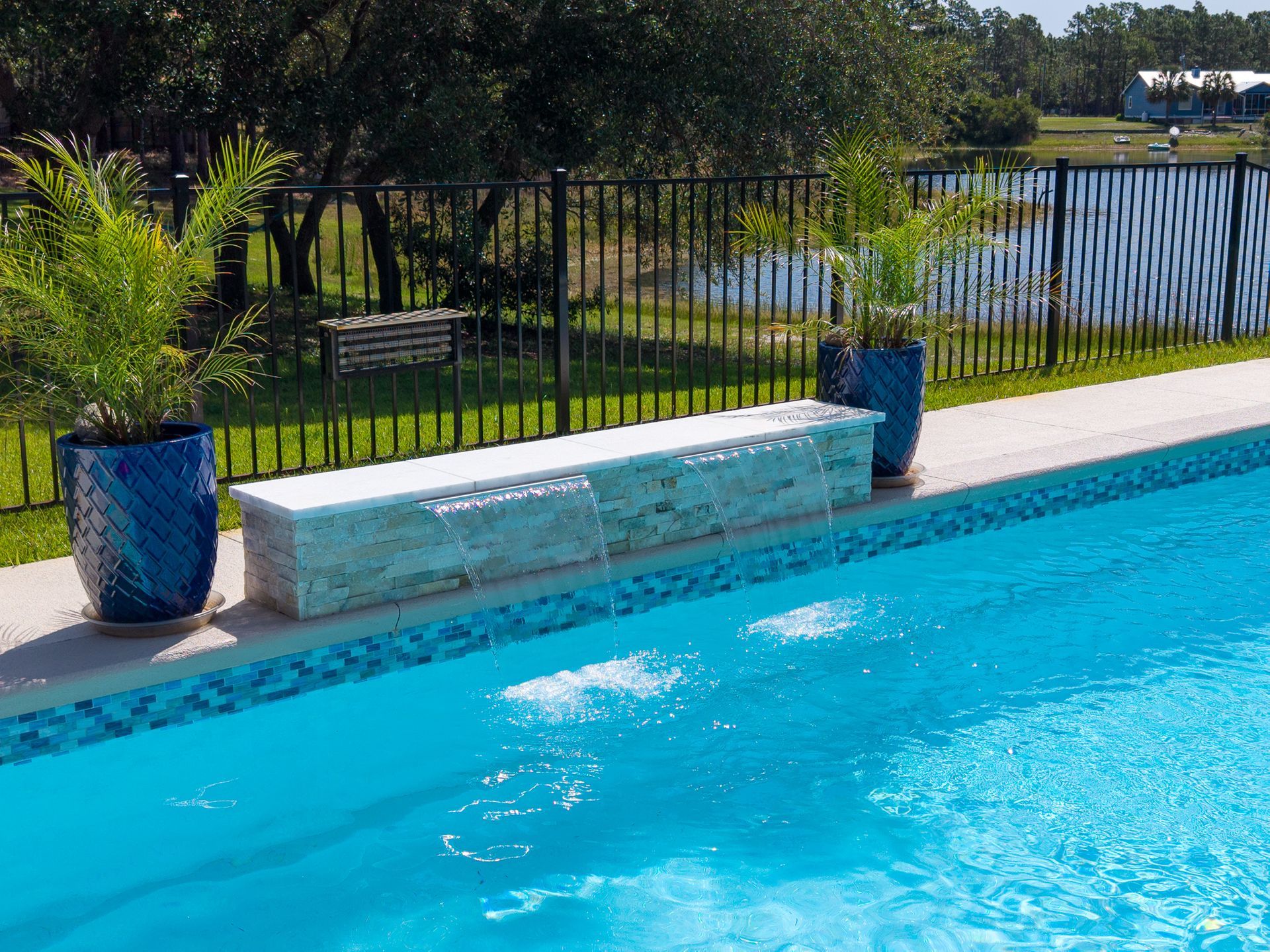Poolside bench beside bright blue swimming pool, with potted plants and a fence by the water.