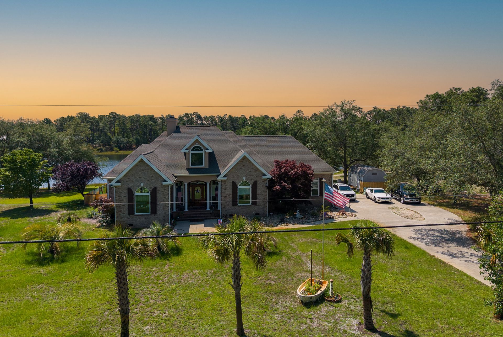 A brick house with a front lawn and palm trees at sunset, with cars parked in the driveway.