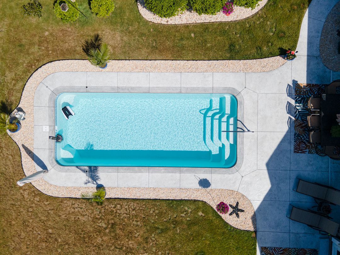Aerial view of a backyard swimming pool with turquoise water, lounge chairs, and surrounding patio and grass.