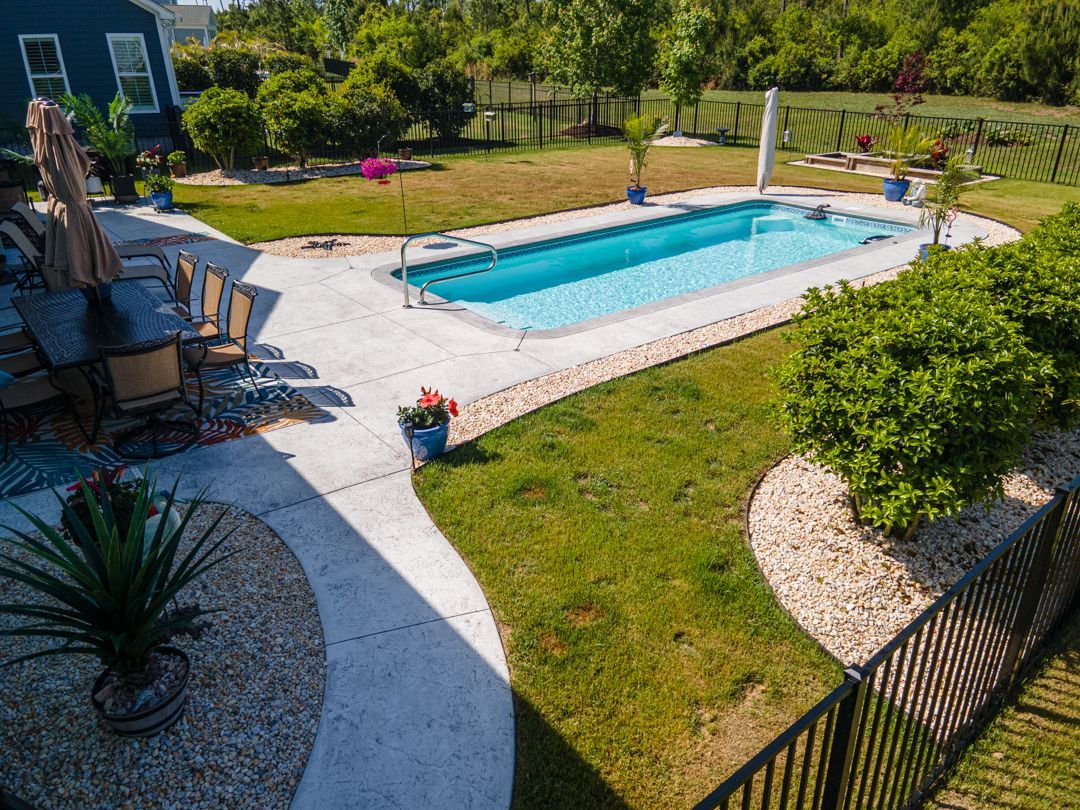 Backyard with a rectangular blue swimming pool, patio chairs, and green lawn bordered by shrubs and a black fence