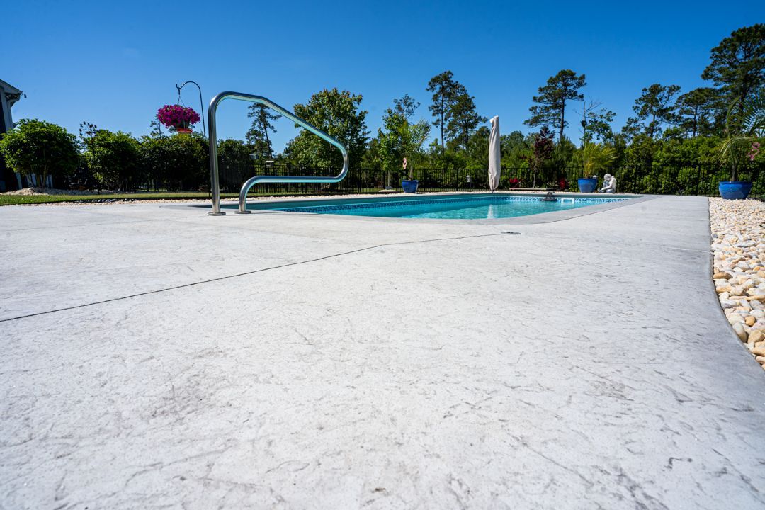 Empty white pool deck beside a turquoise pool with a basketball hoop and palm trees under a clear blue sky