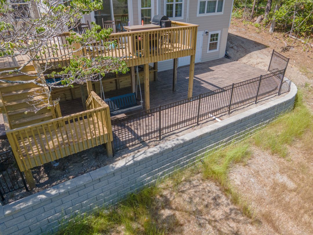 Raised wooden deck with stairs and a stone retaining wall beside a house in a backyard