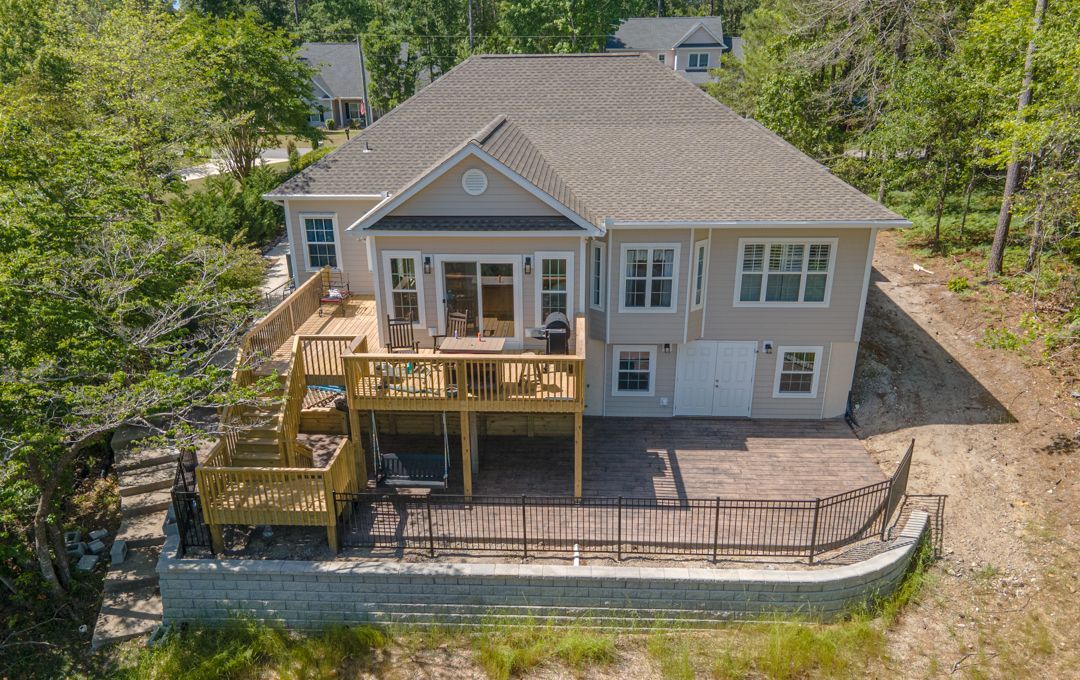 Aerial view of a gray house with a large wooden deck and patio in a wooded yard.