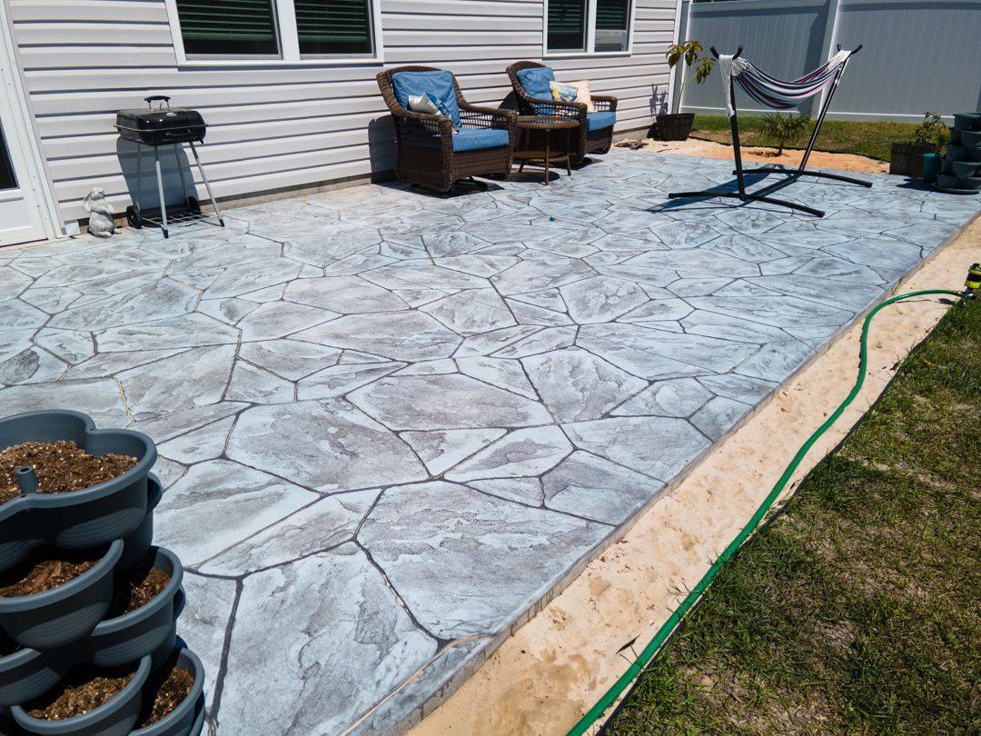 Stamped concrete patio beside a house, with outdoor chairs, grill, and planters along the wall.