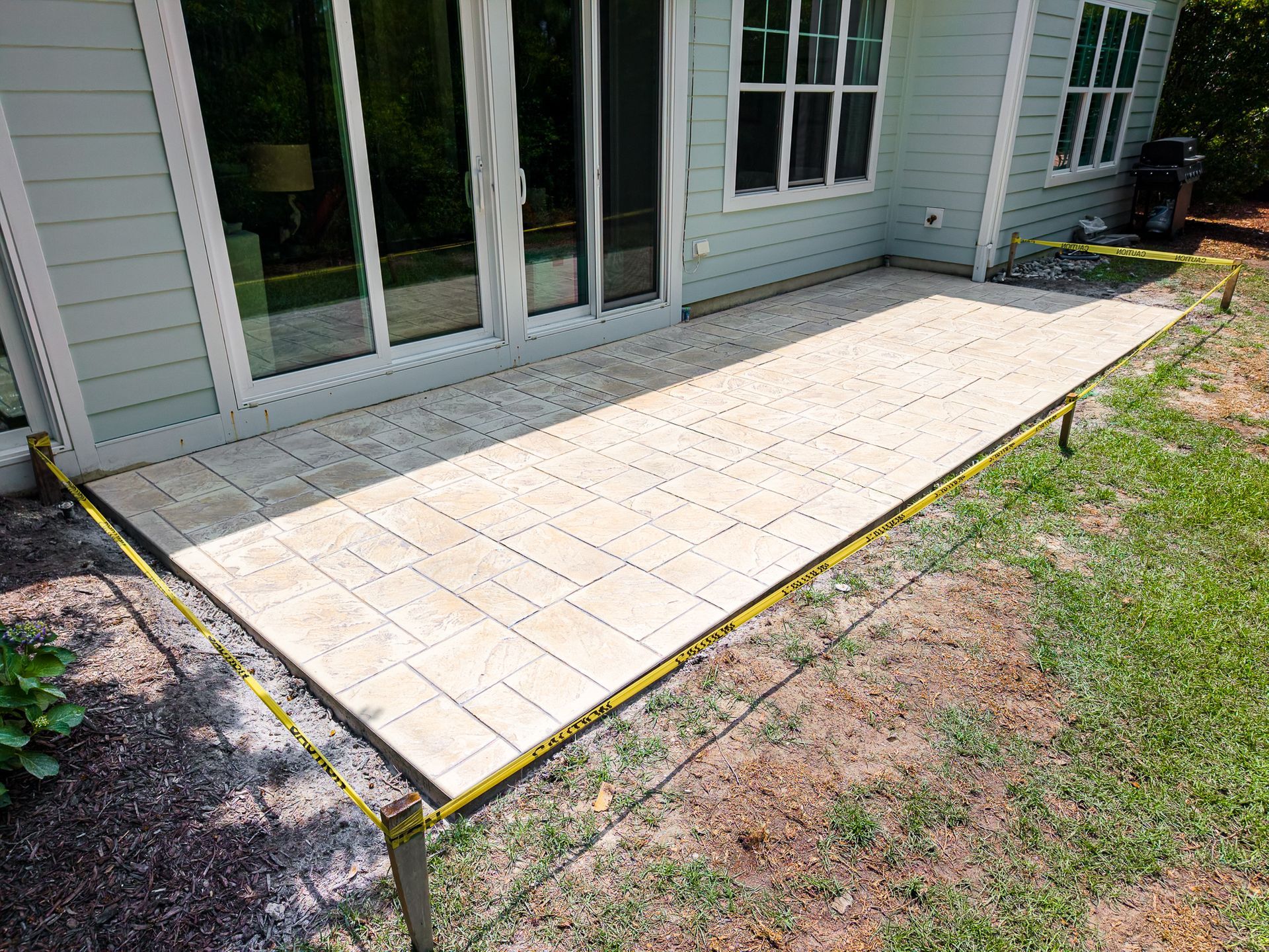 Paver patio beside sliding glass doors, edged with yellow tape, next to a grassy yard.