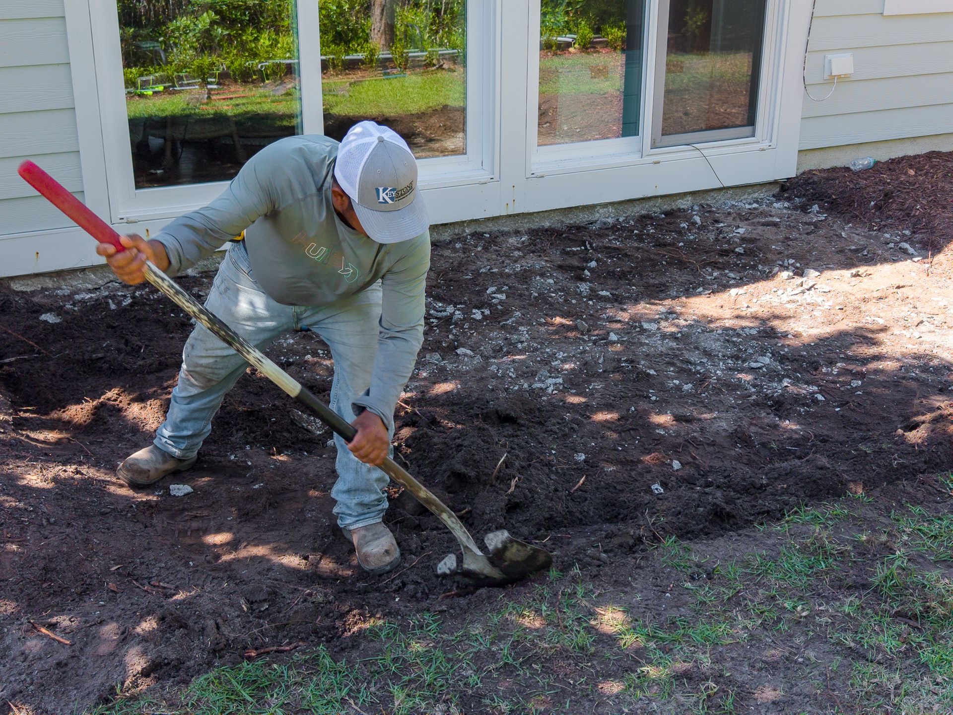 Person raking dark mulch near a house with a red-handled tool