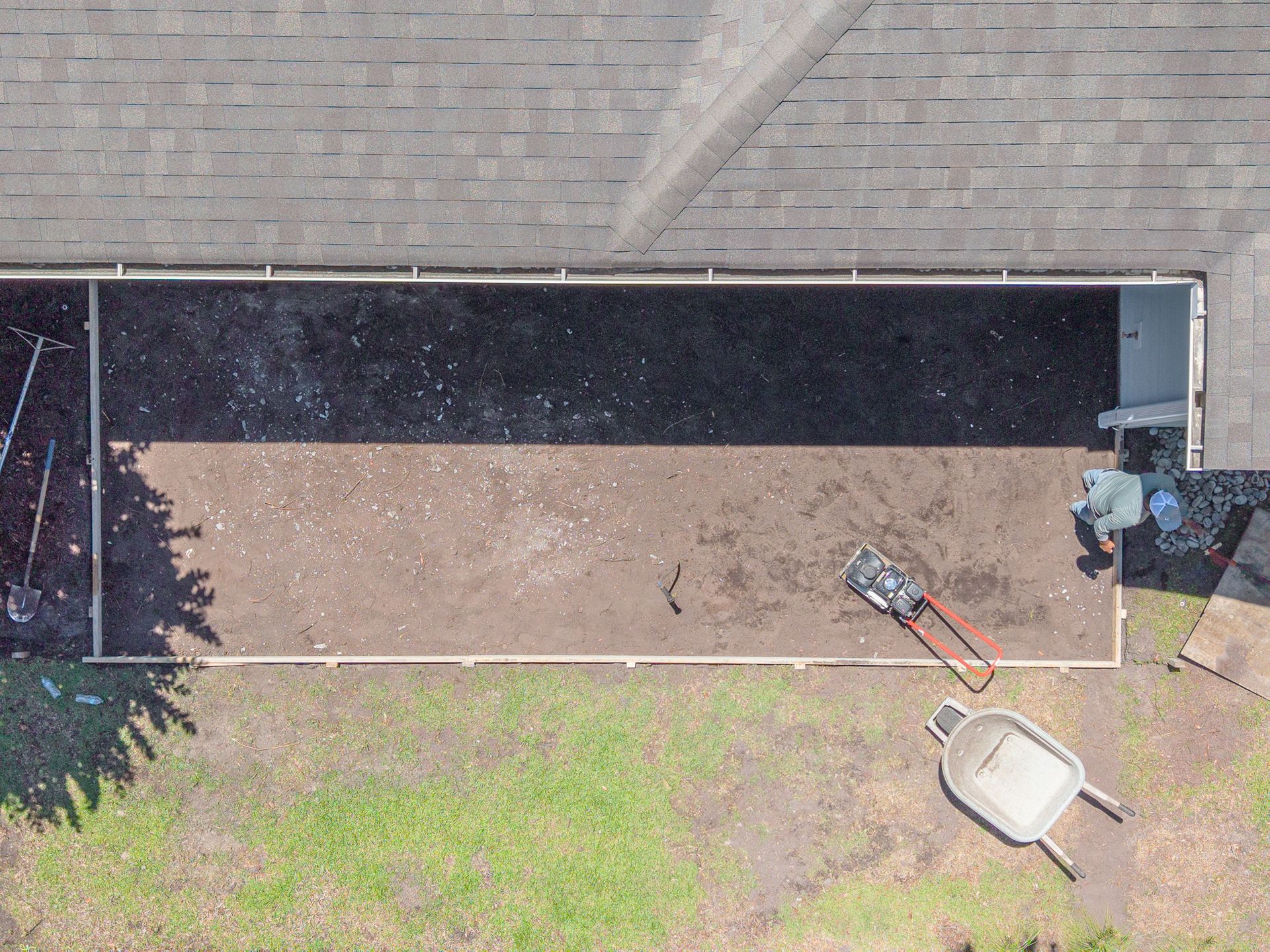 Overhead view of a backyard with a brick wall, a long dirt strip, and a wheelbarrow near a lawn.