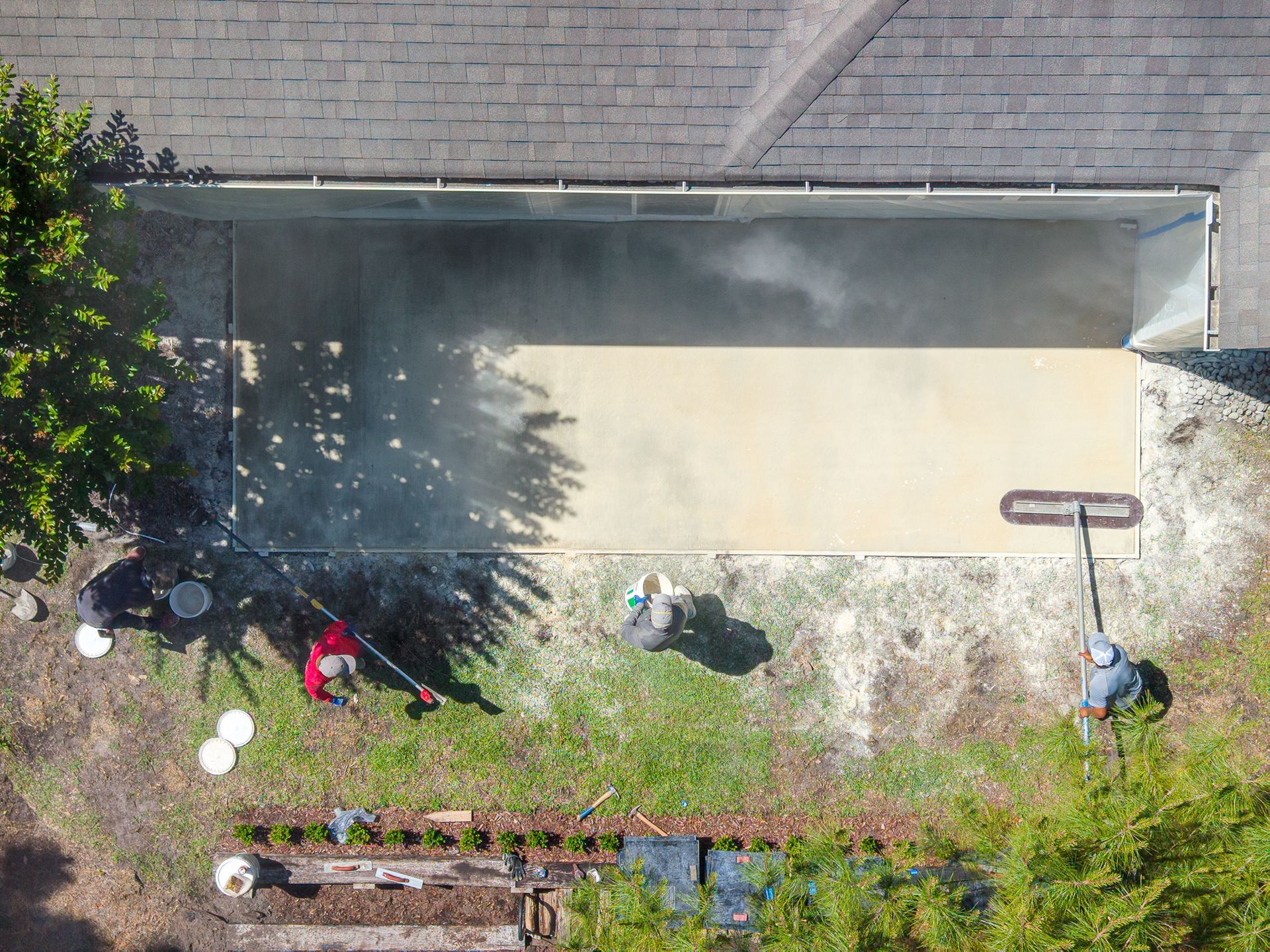 Aerial view of a backyard with a house wall, patio, bushes, and garden beds along the fence.