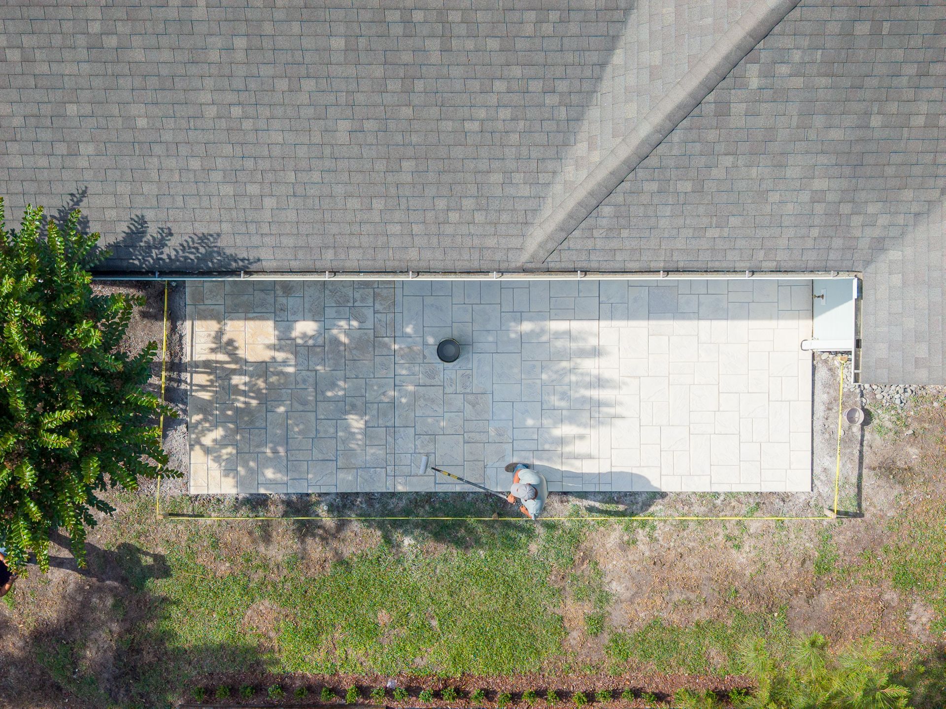 Aerial view of a narrow concrete patio beside a gray roof, bordered by grass and a tree.