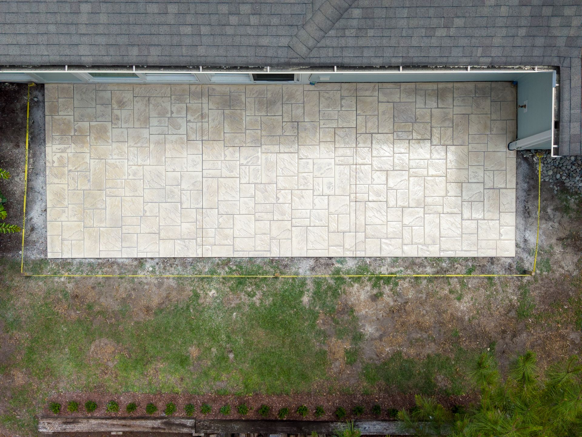 Overhead view of a small stone-paved patio beside a building, bordered by grass and garden edges.