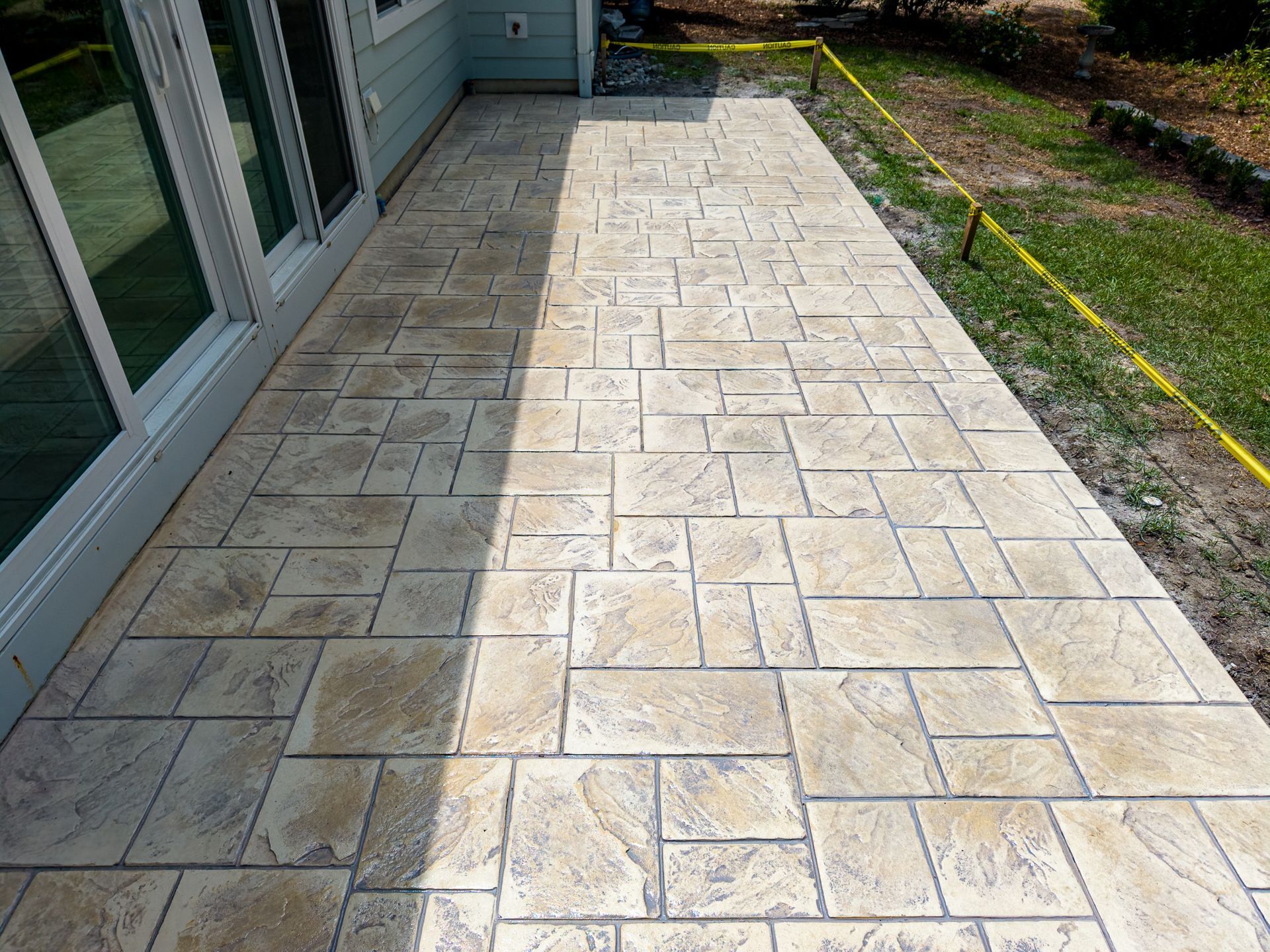 Sunlit tiled patio beside a house, with sliding glass doors and a grassy yard alongside.