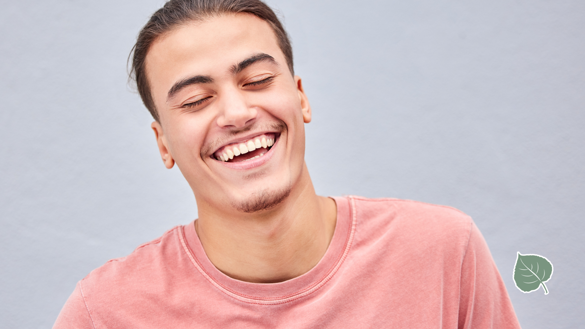 Man with brown hair in a bun, laughing and wearing a pink shirt against a grey backdrop.
