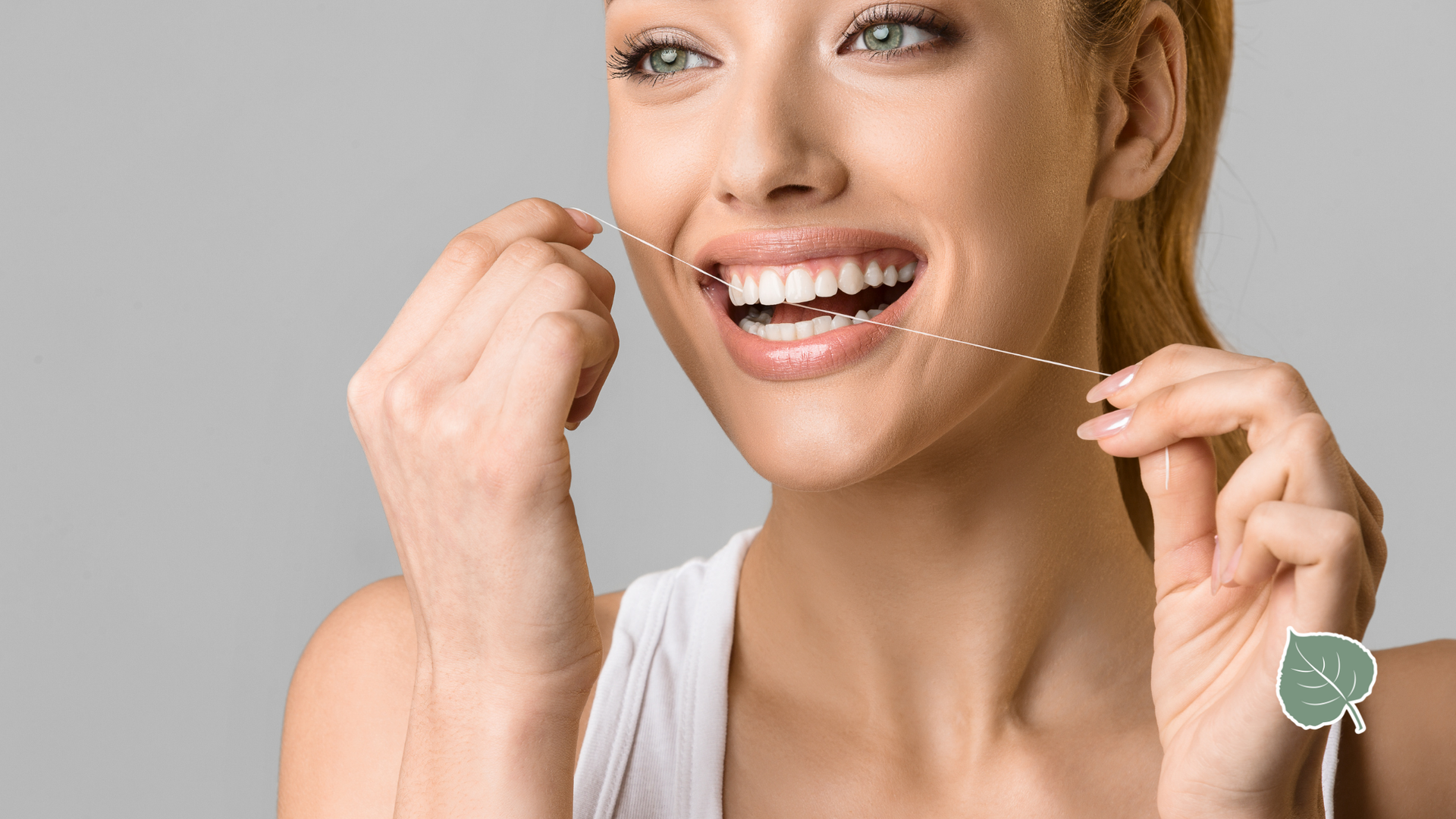 Woman flossing her teeth, smiling widely. White tank top, gray background.