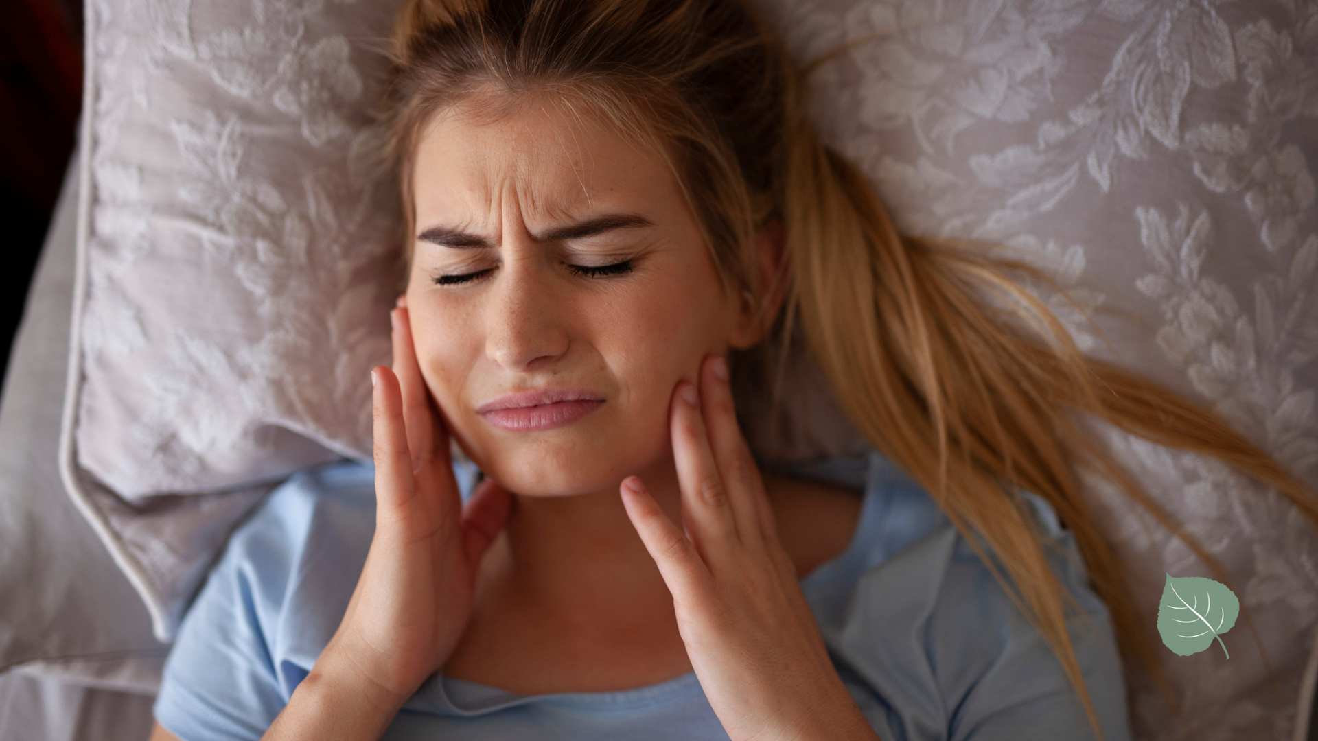 Woman in bed, clutching cheeks with pained expression, likely experiencing jaw or tooth pain.
