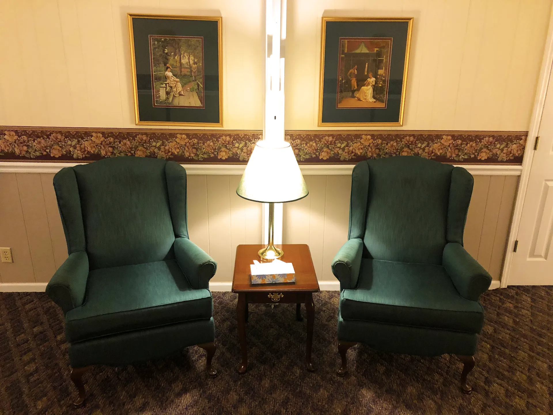 Two green armchairs face each other, with a small table and lamp in between. Artwork hangs on the wall.