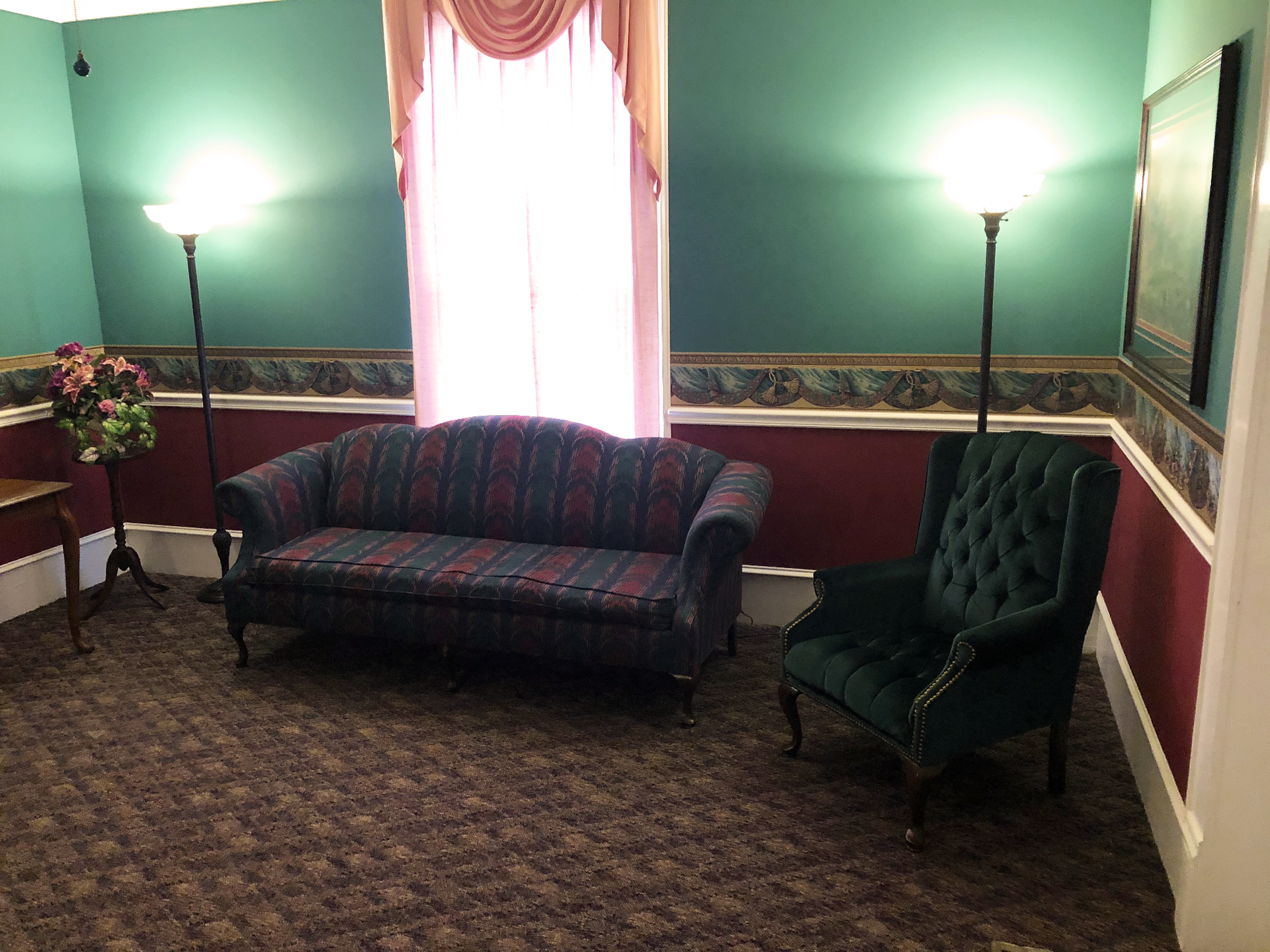 A room with a sofa, chair, and lamps; red and green walls, with a window and carpet.