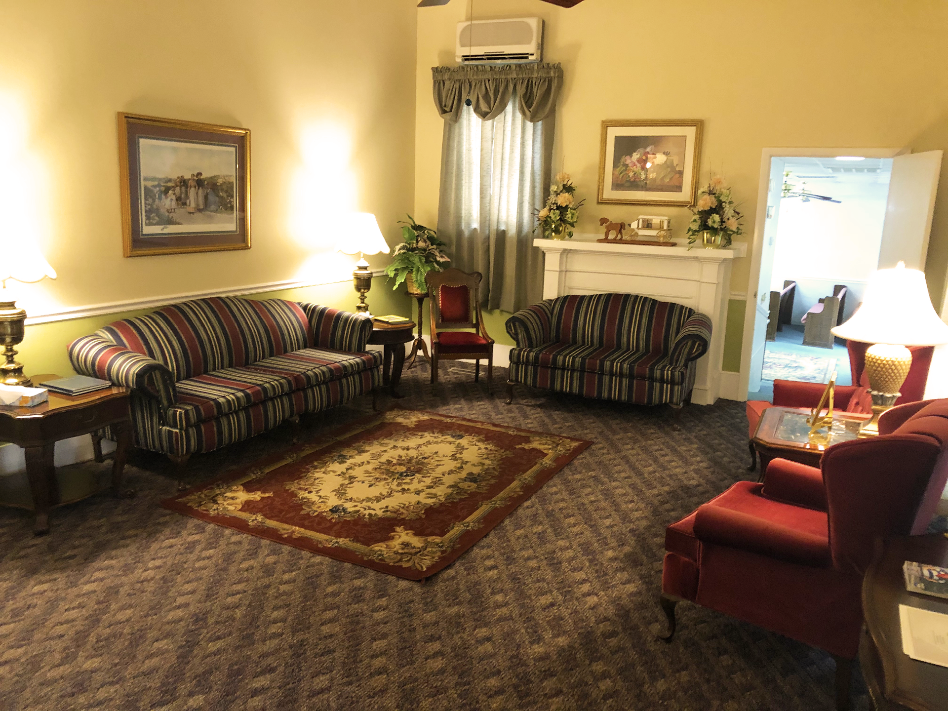 A furnished waiting room with two patterned sofas, red chairs, carpet, and artwork on the walls.
