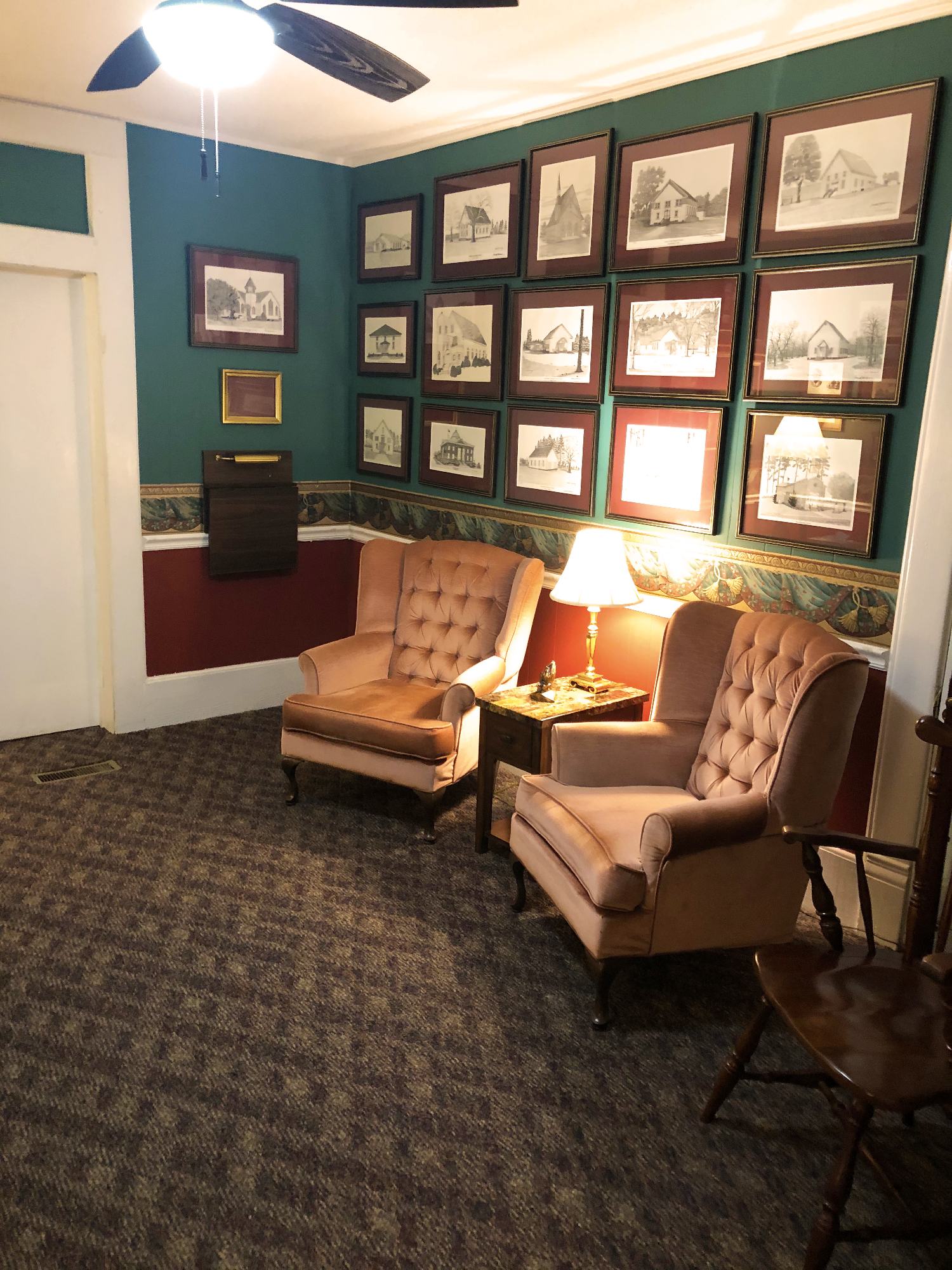 Two armchairs and a small table with a lamp, set against a wall with framed pictures. Green wall with pictures and carpeted floor.