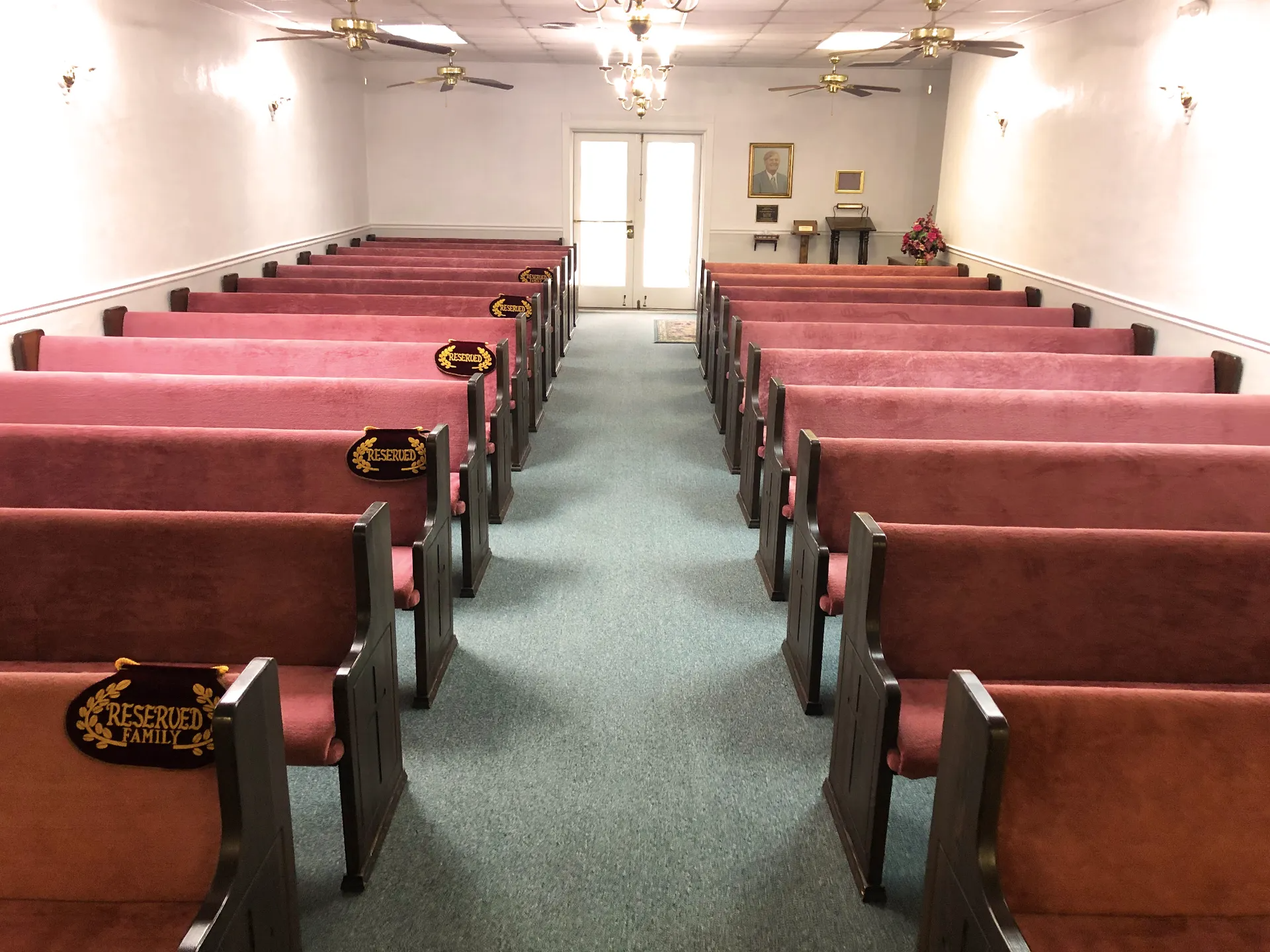 Rows of empty pews with burgundy cushions in a chapel. Door at end, ceiling lights.