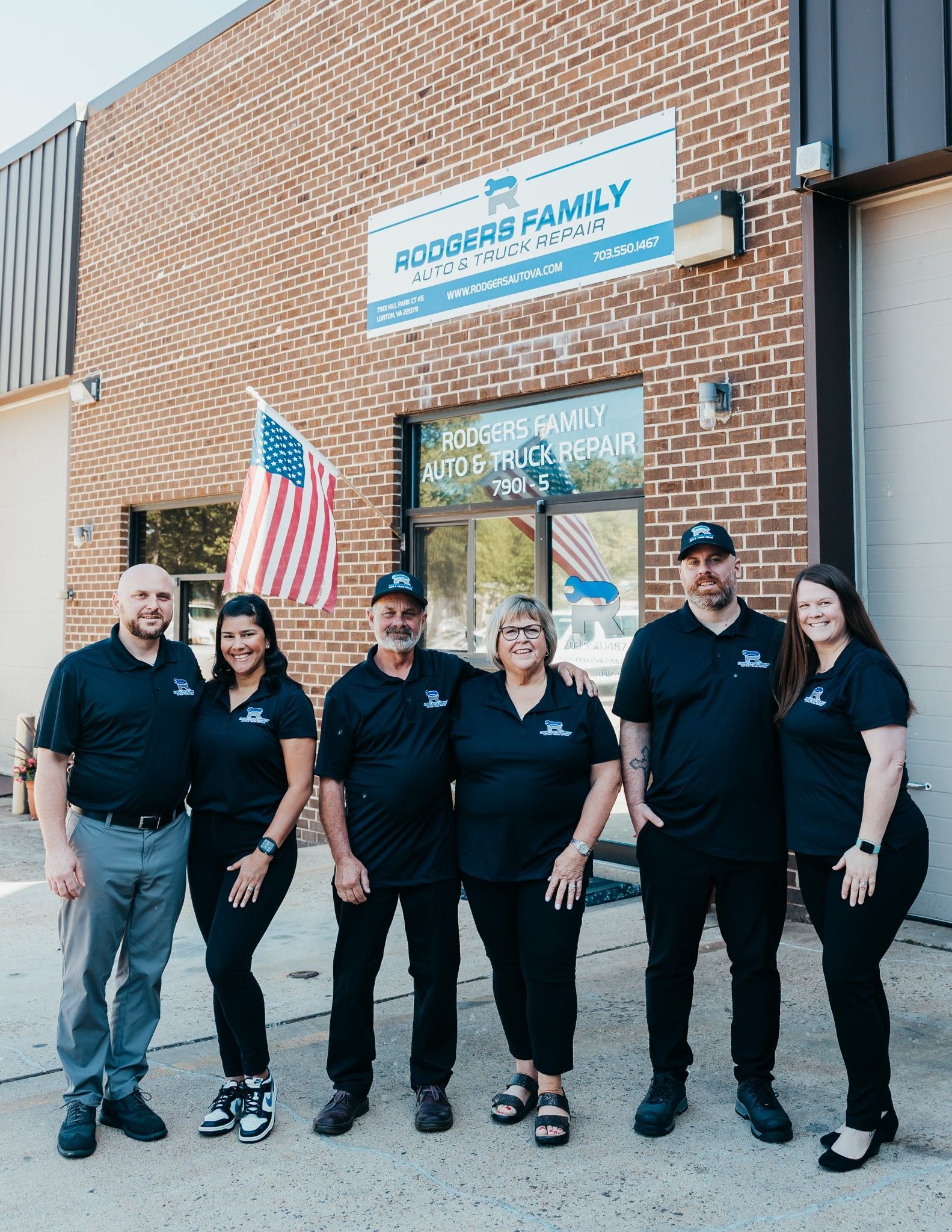 Six people in black shirts stand outside a brick building with an American flag and a sign above the door.