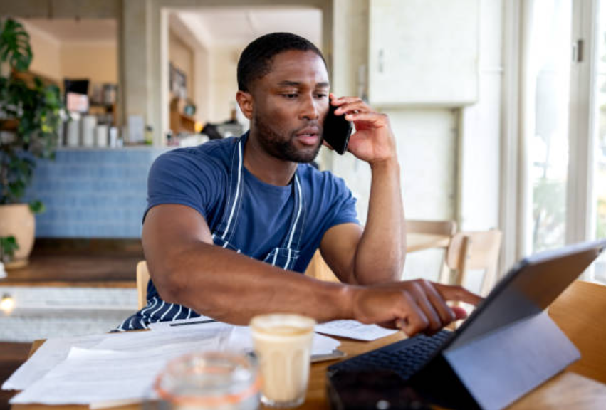 Man in blue shirt and striped apron on phone, using tablet at table with coffee, papers, and window in background.