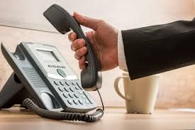 Hand holding a black telephone receiver, reaching for a desk phone in an office setting.