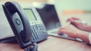 Black office phone on desk with laptop and hands typing in background.
