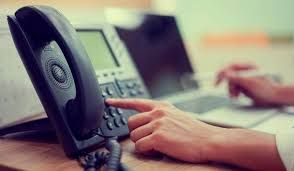 Hand pressing a button on a black office phone, laptop in background, blurry setting.