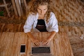 Woman with curly hair types on a laptop at a wooden table, phone nearby.