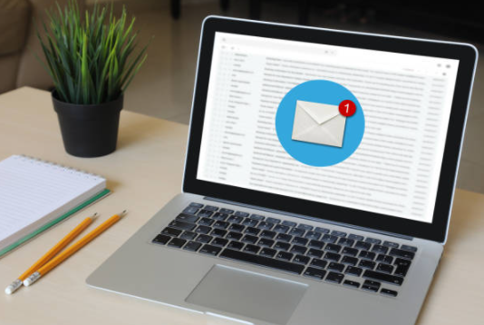 Laptop displaying an email inbox, with a blue notification icon. A plant, notebook, and pencils sit nearby.