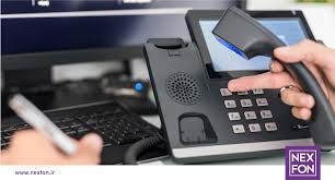 A hand dials a number on a black office phone, near a keyboard and computer screen.
