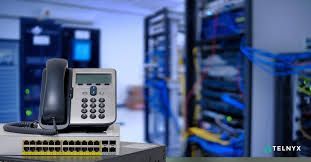 VoIP phone on top of a network switch in a server room with blue cables and servers.