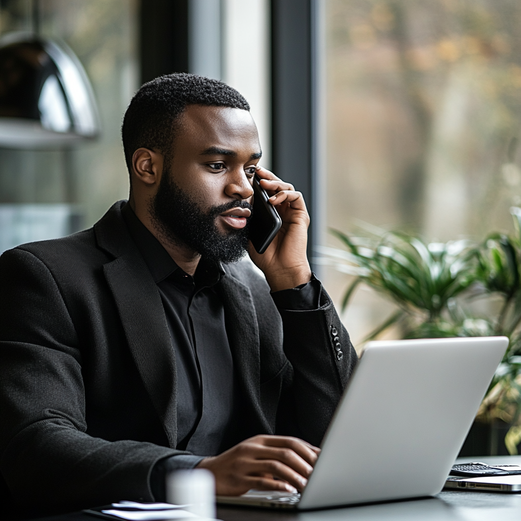Man in a black suit, on the phone, working on a laptop at a table near a window.