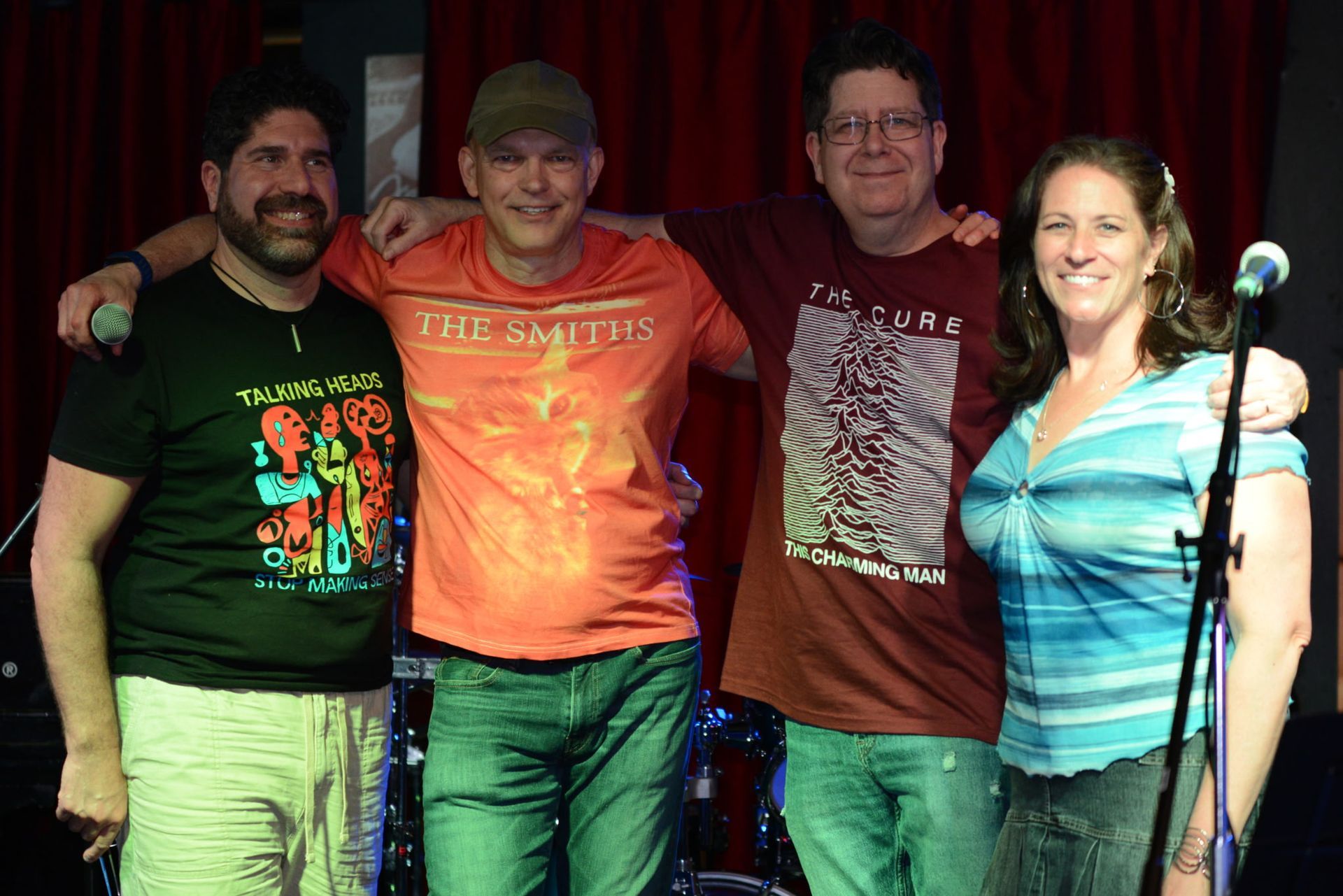 Four people standing together on a small stage with a red curtain backdrop, smiling beside a microphone.