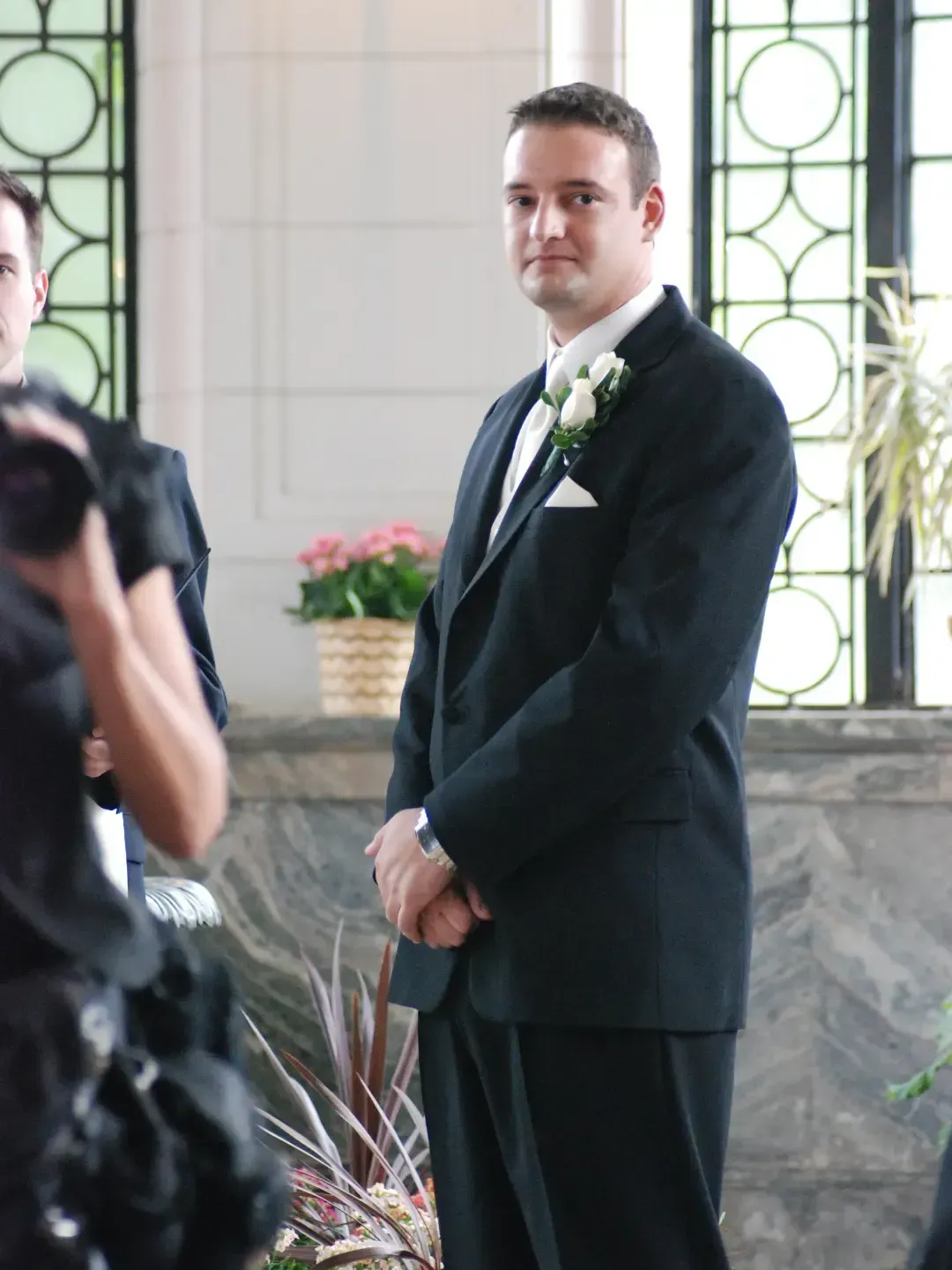 Man in a black suit stands indoors beside a photographer, with floral décor and windows behind him