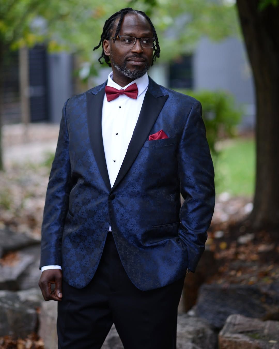 Man in navy tuxedo with red bow tie and pocket square standing outdoors on a path