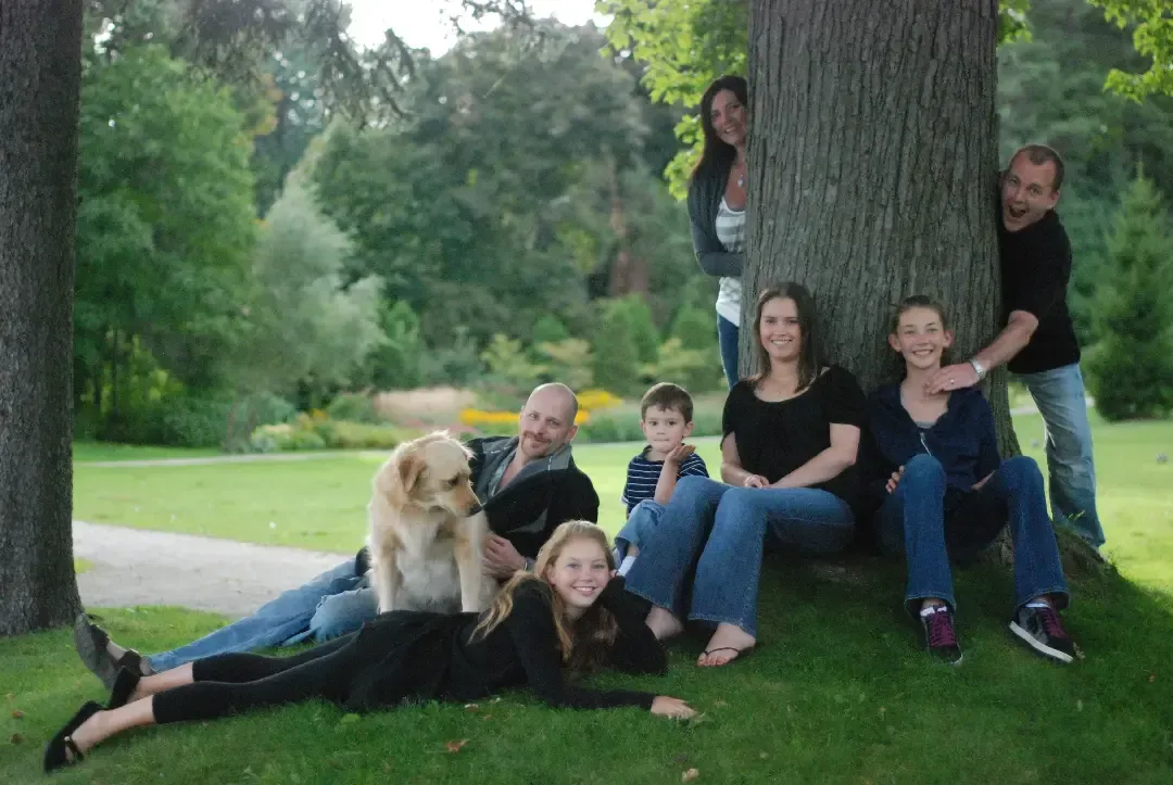Group of people posing on grass beside a large tree in a park, with a wooded background.