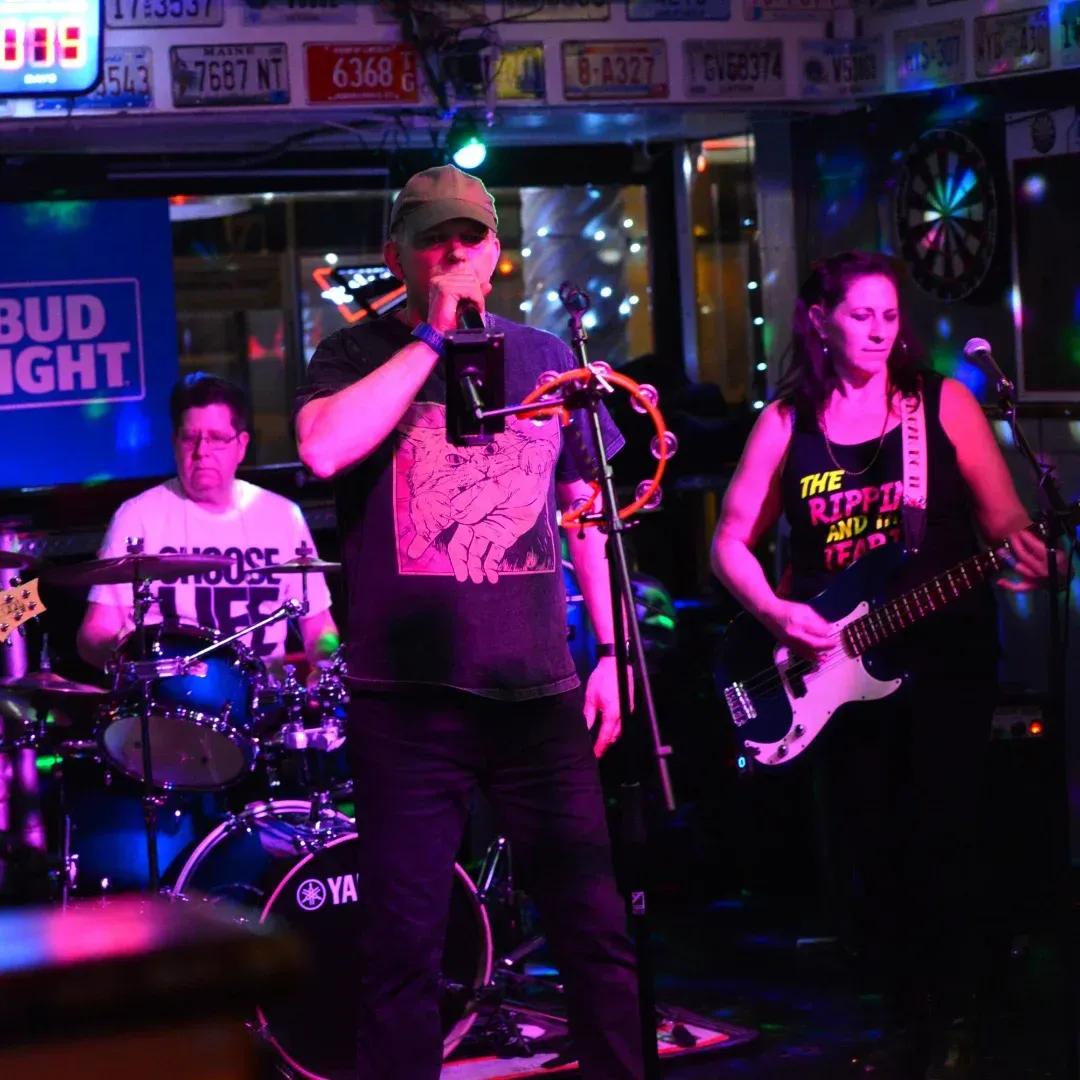 Live band performing in a neon-lit bar, with a singer, drummer, and bassist on stage.