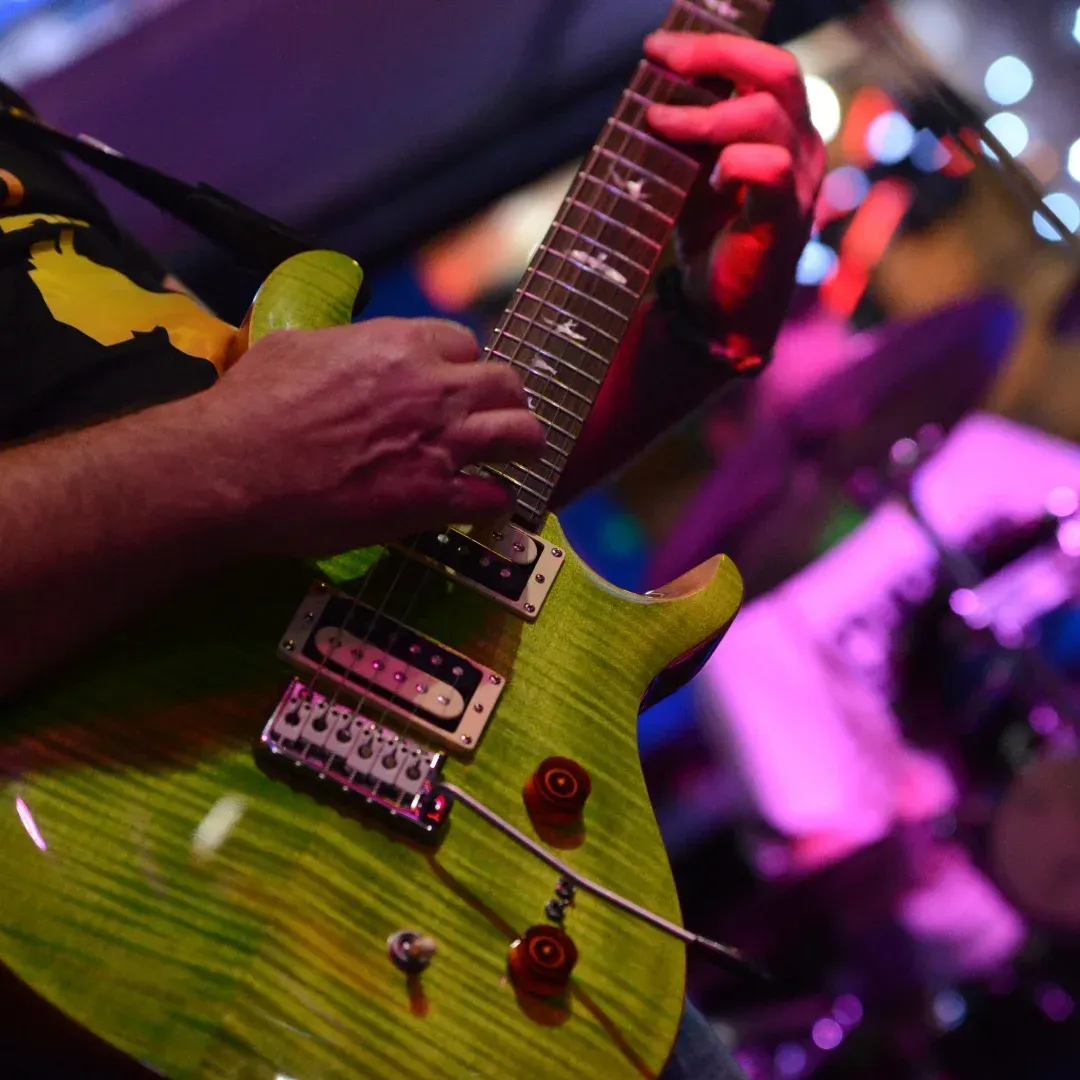 Close-up of hands playing a green electric guitar under purple stage lights.