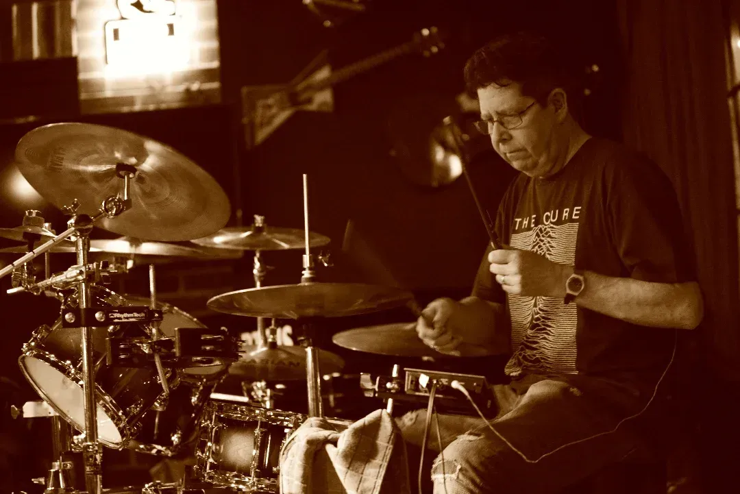 Drummer seated at a drum kit under warm sepia lighting, mid-performance in a dim venue