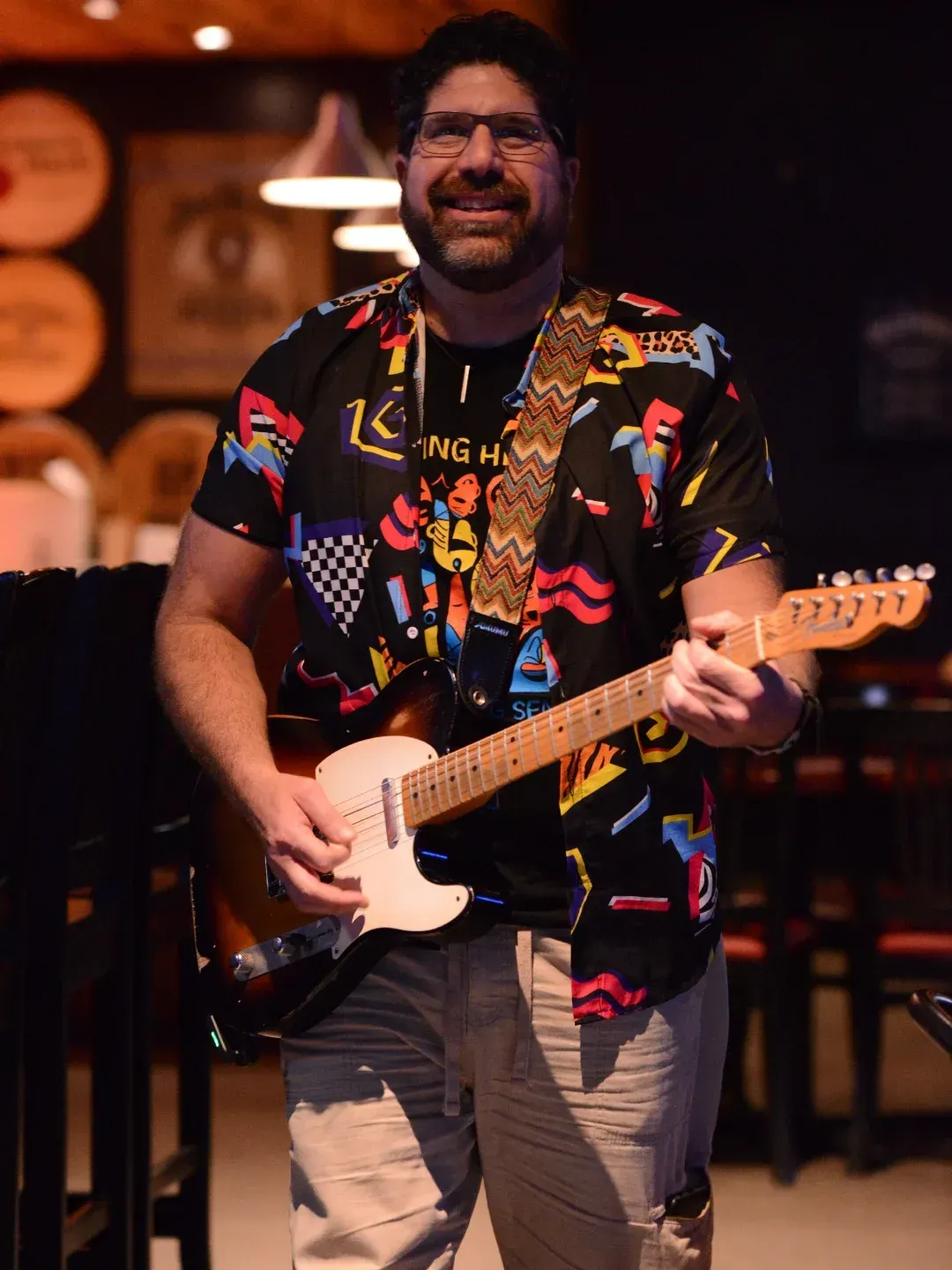 Smiling musician playing electric guitar on a dimly lit stage, wearing a colorful shirt.